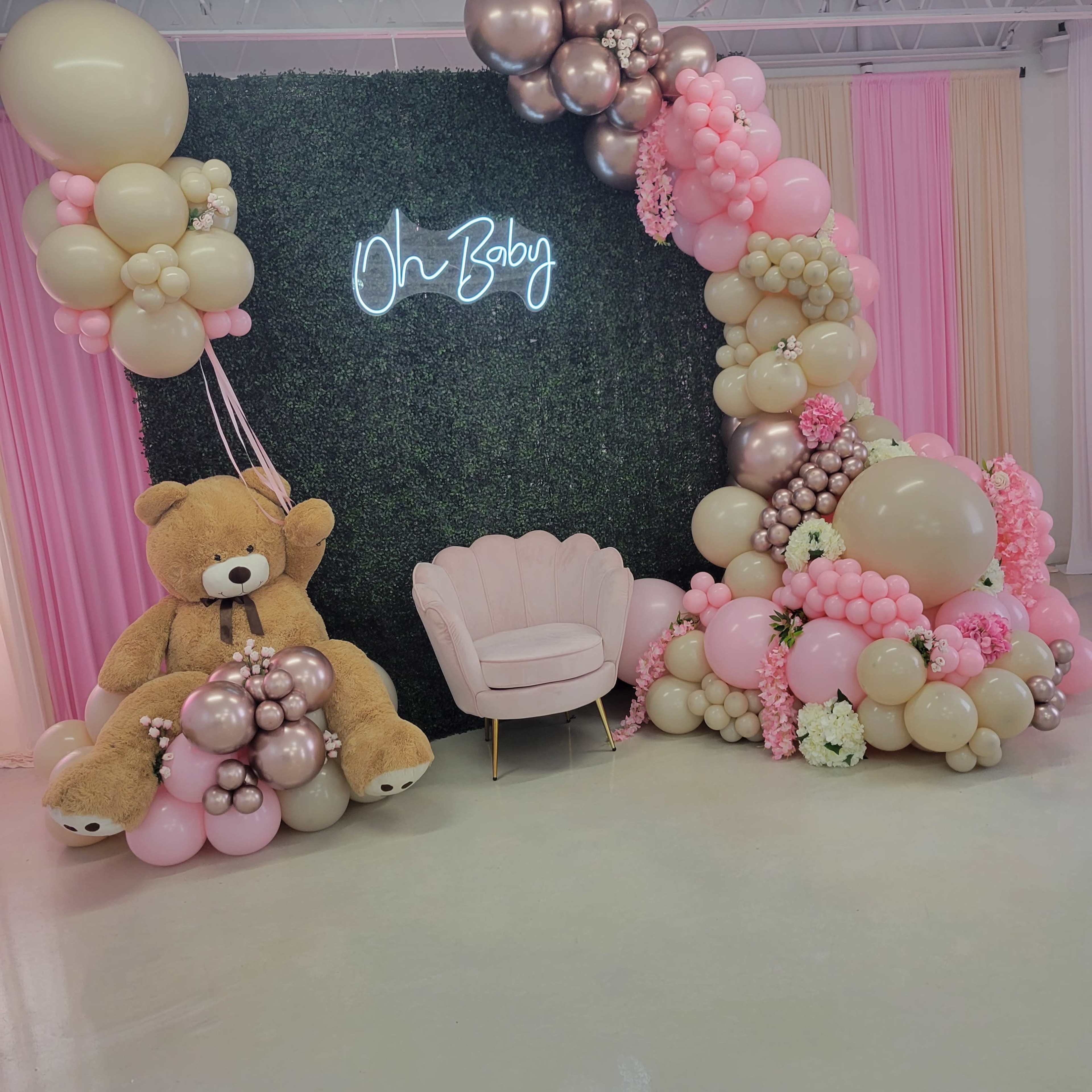 A decorated space with a green floral backdrop, pink and gold balloons, a large teddy bear, and a blush-colored chair, all set for a celebration with the phrase "Oh Baby" illuminated.