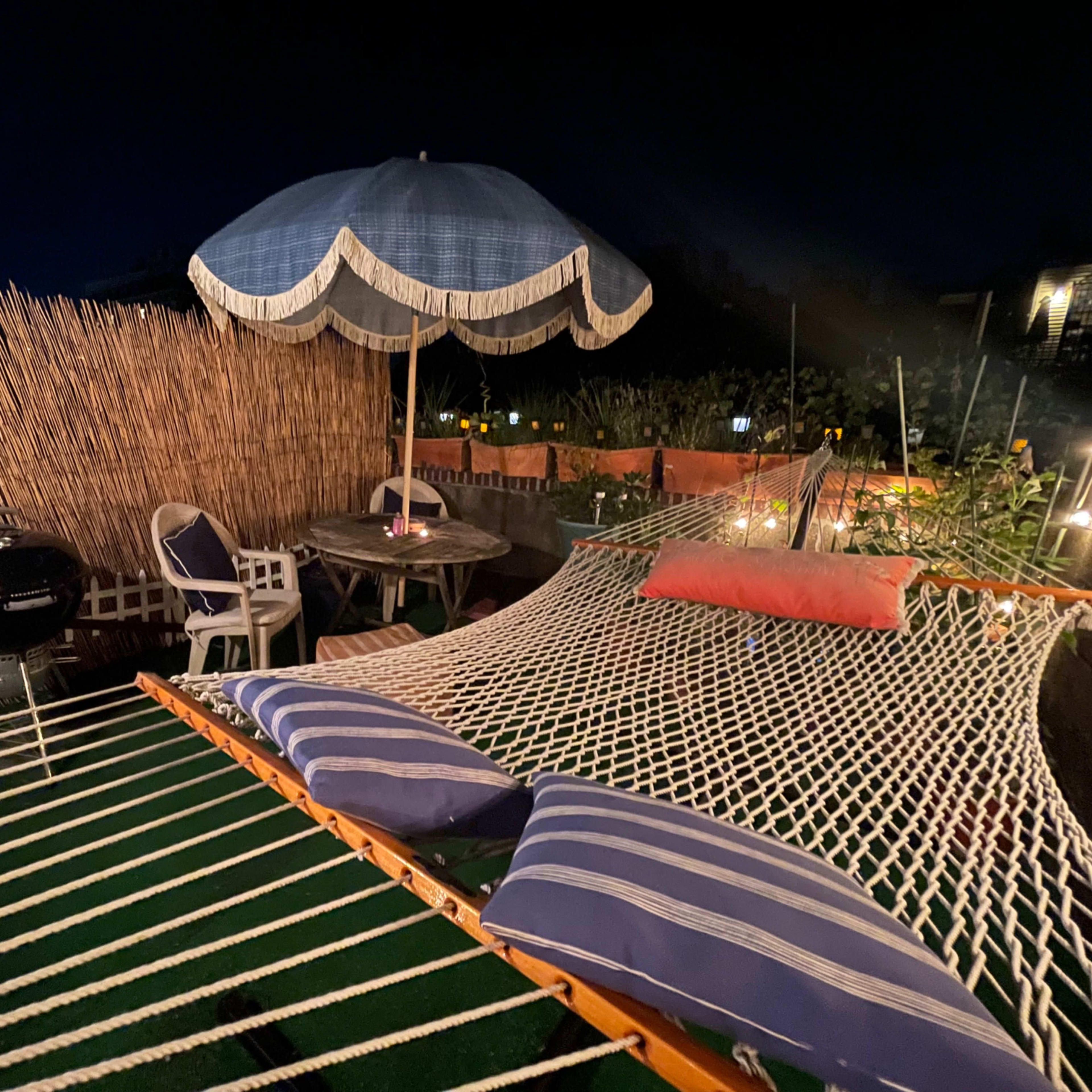 A hammock with striped cushions is set up under a blue umbrella in a dimly lit outdoor space surrounded by a bamboo fence and a small table with chairs.