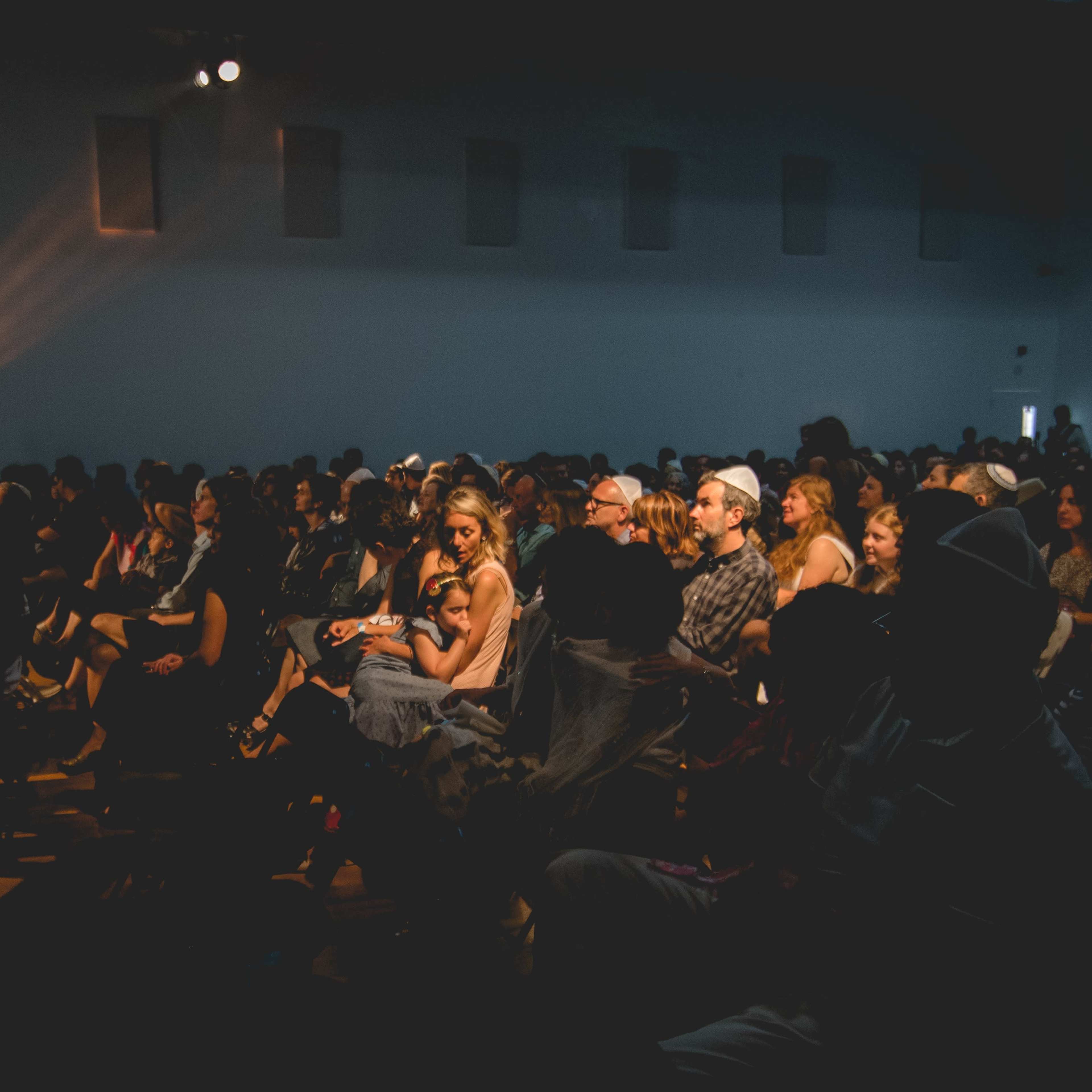 A large audience is gathered in a dimly lit indoor venue, attentively facing the front stage.