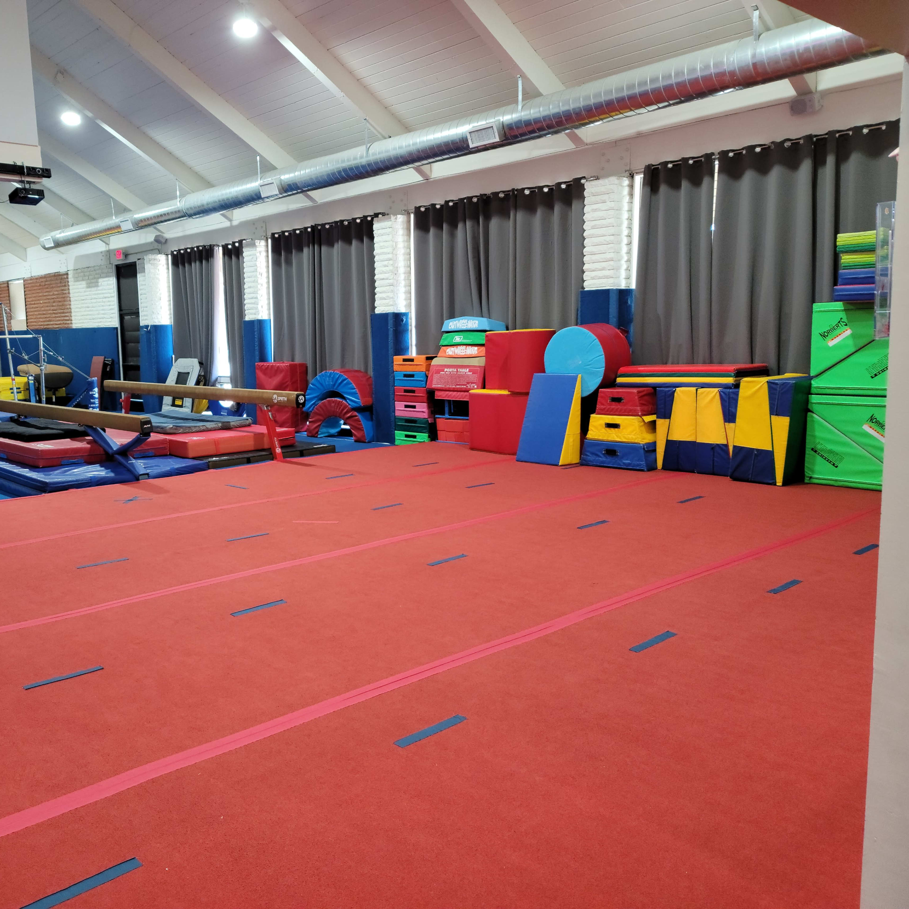 A gymnastics gym with red carpet flooring, various training equipment, and colorful mats arranged along the walls.