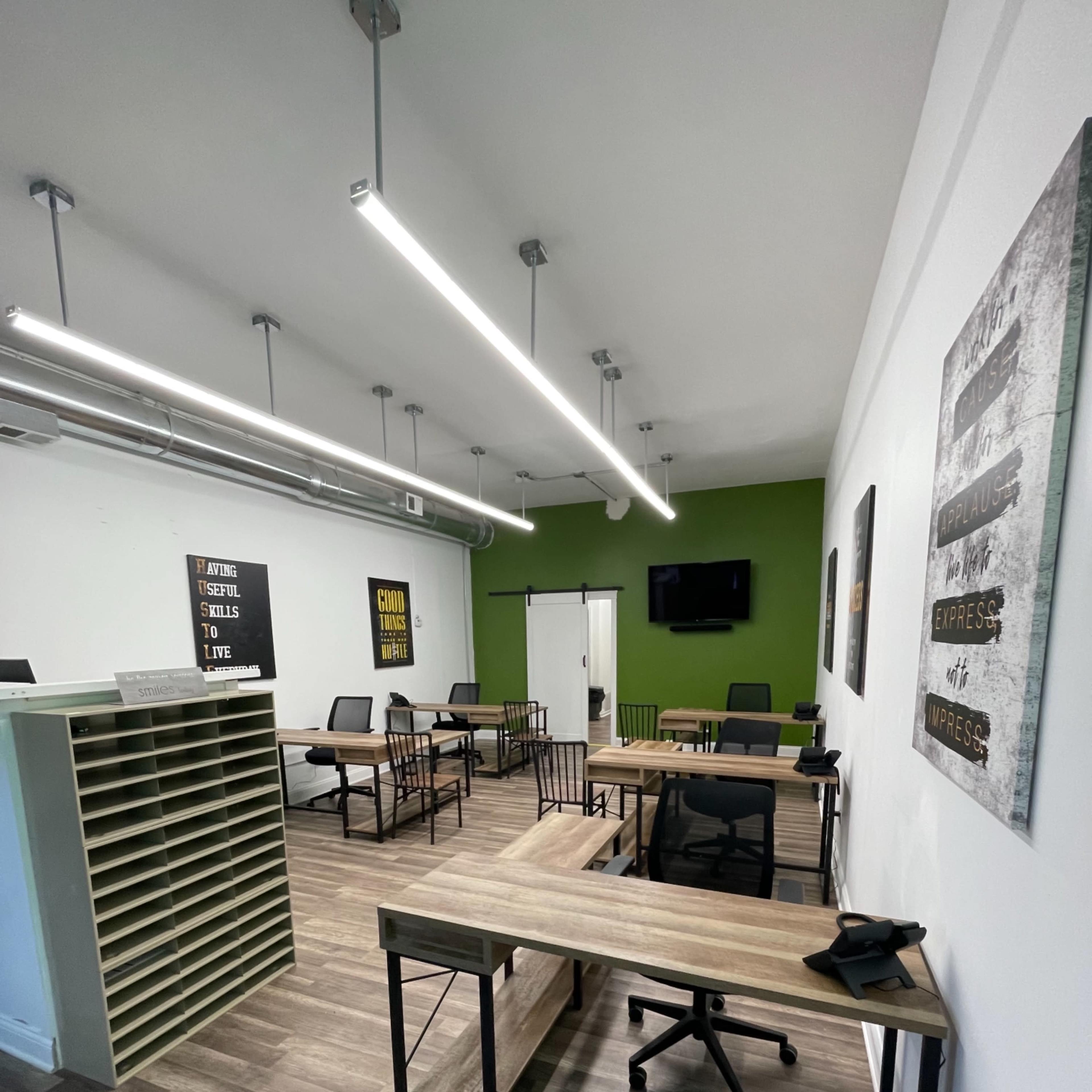 A modern office space with several desks, chairs, and a green accent wall, featuring both shelving and a television mounted on the wall.