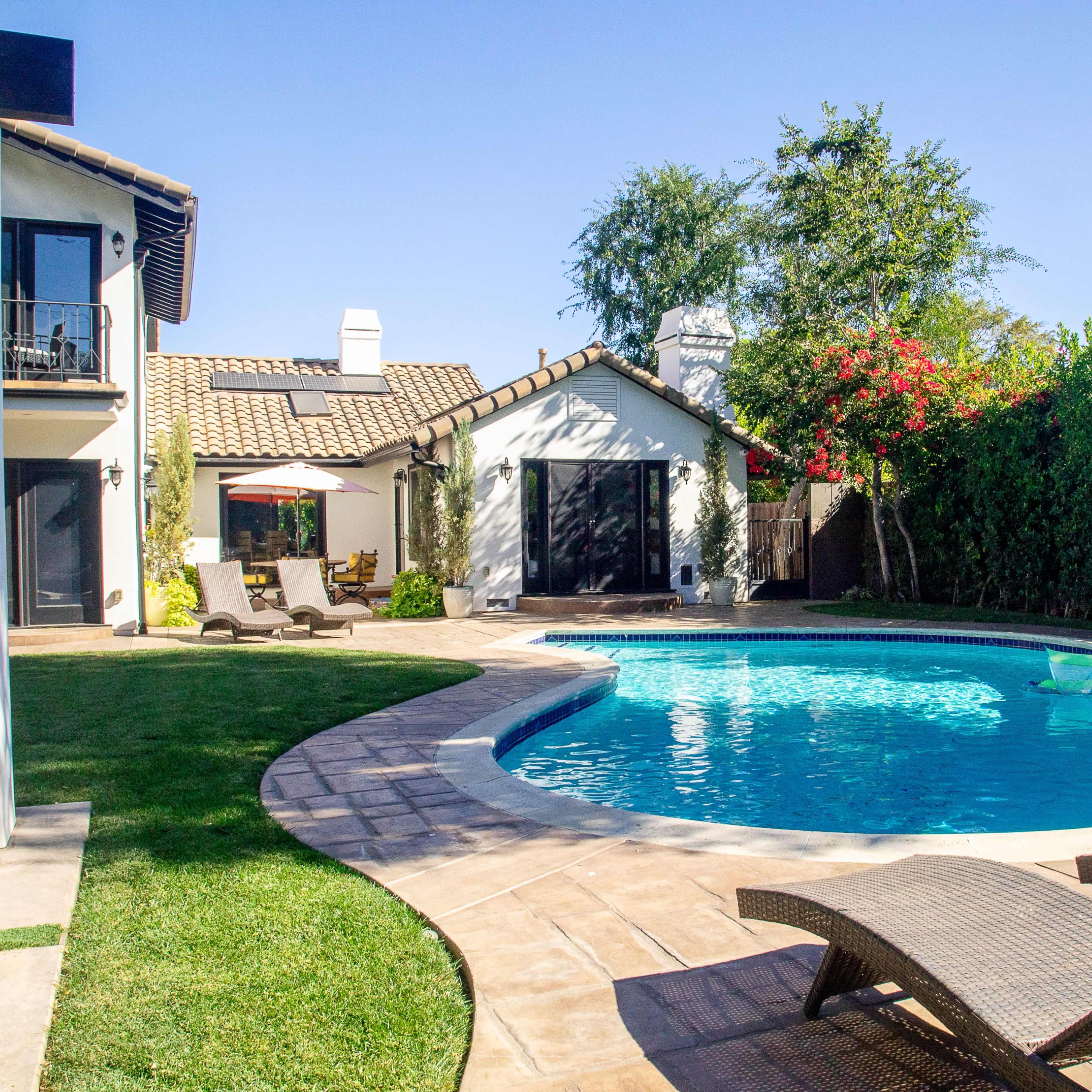A backyard features a curved swimming pool surrounded by lounge chairs, with a house and garden in the background.