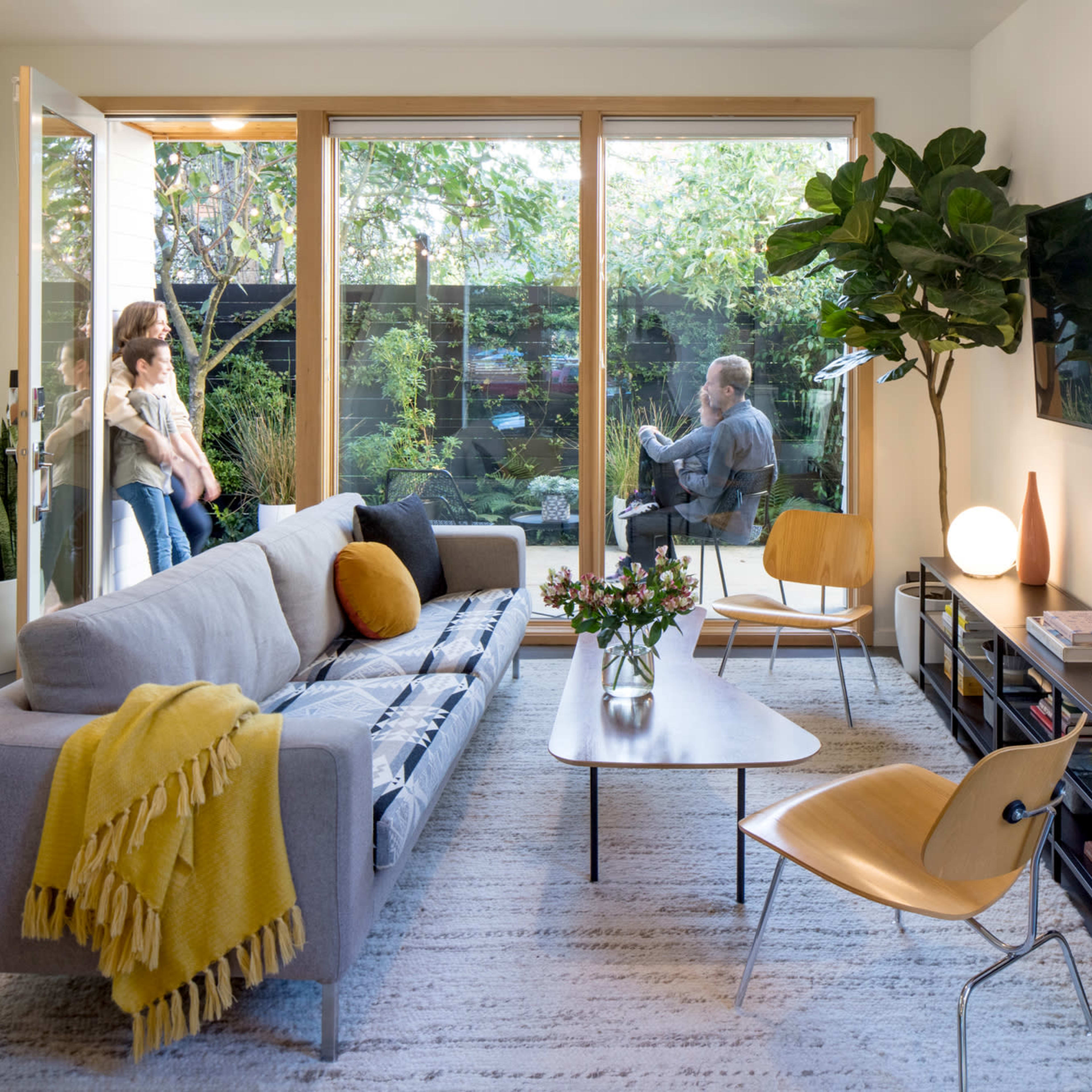 A modern living room features a gray sofa with a yellow throw, a coffee table, and a large window opening to a garden, while a person sits outside and two others stand inside.