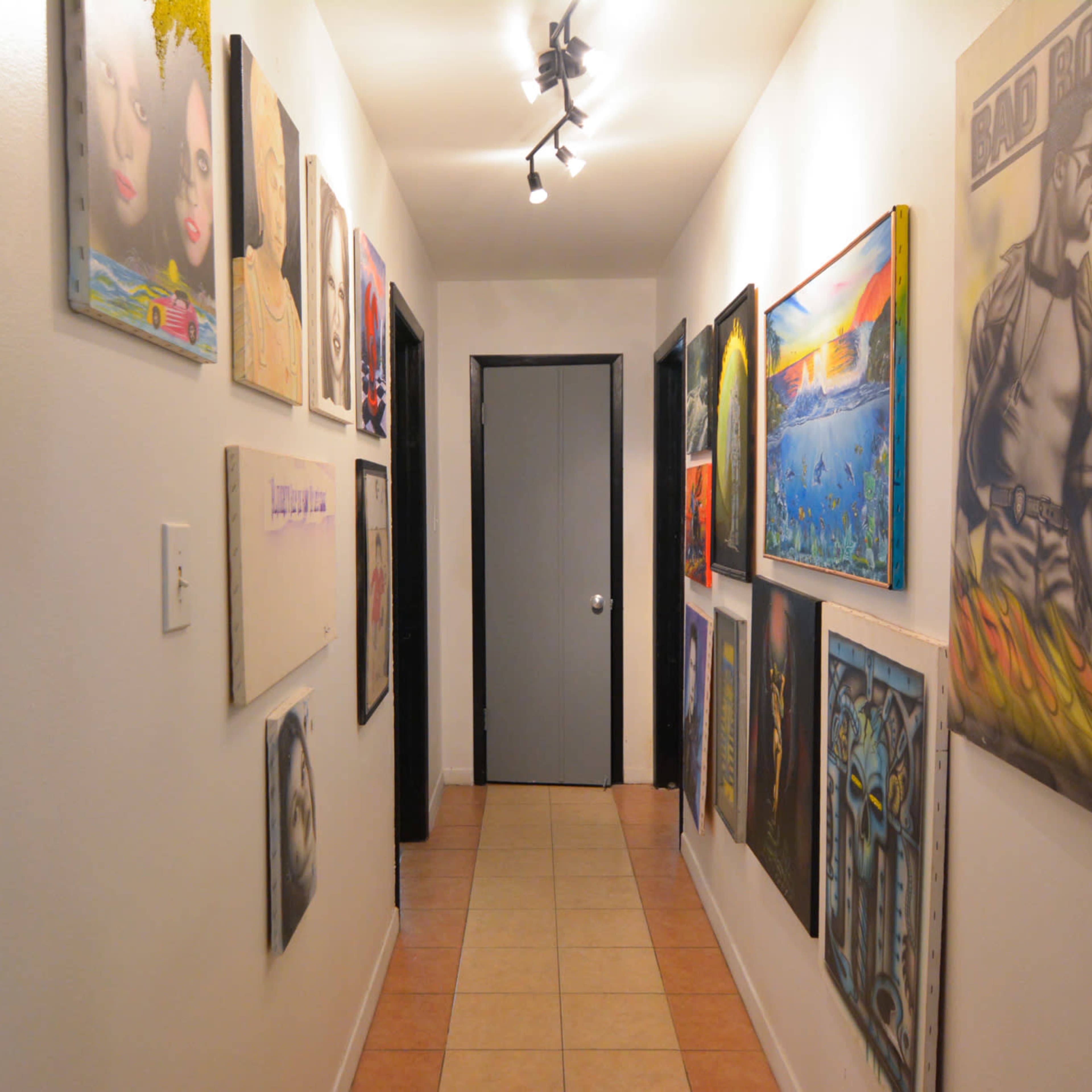 A narrow hallway features framed artworks hung on both sides, leading to a closed door at the end.