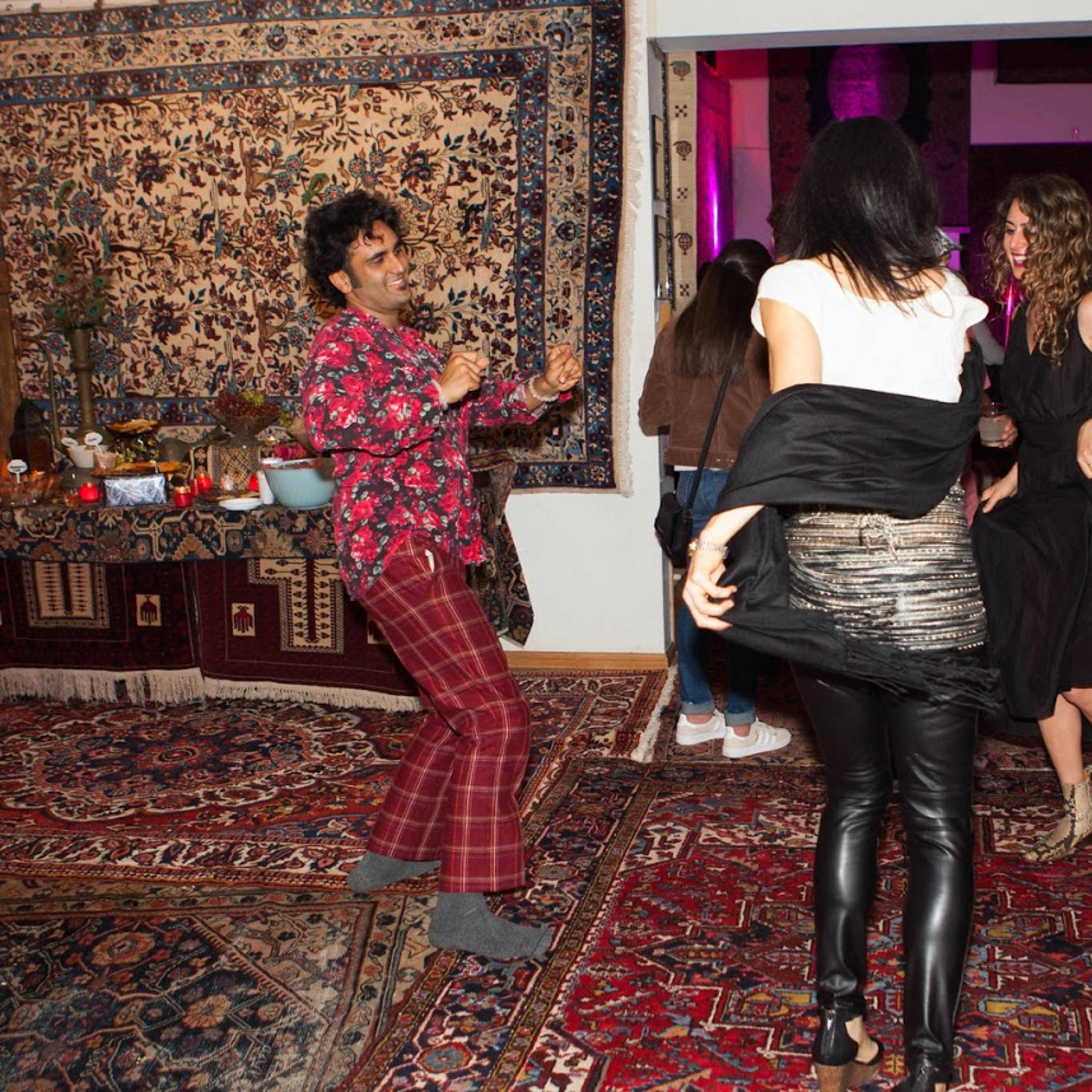 A group of people dance at a festive gathering surrounded by colorful rugs and intricate wall hangings.