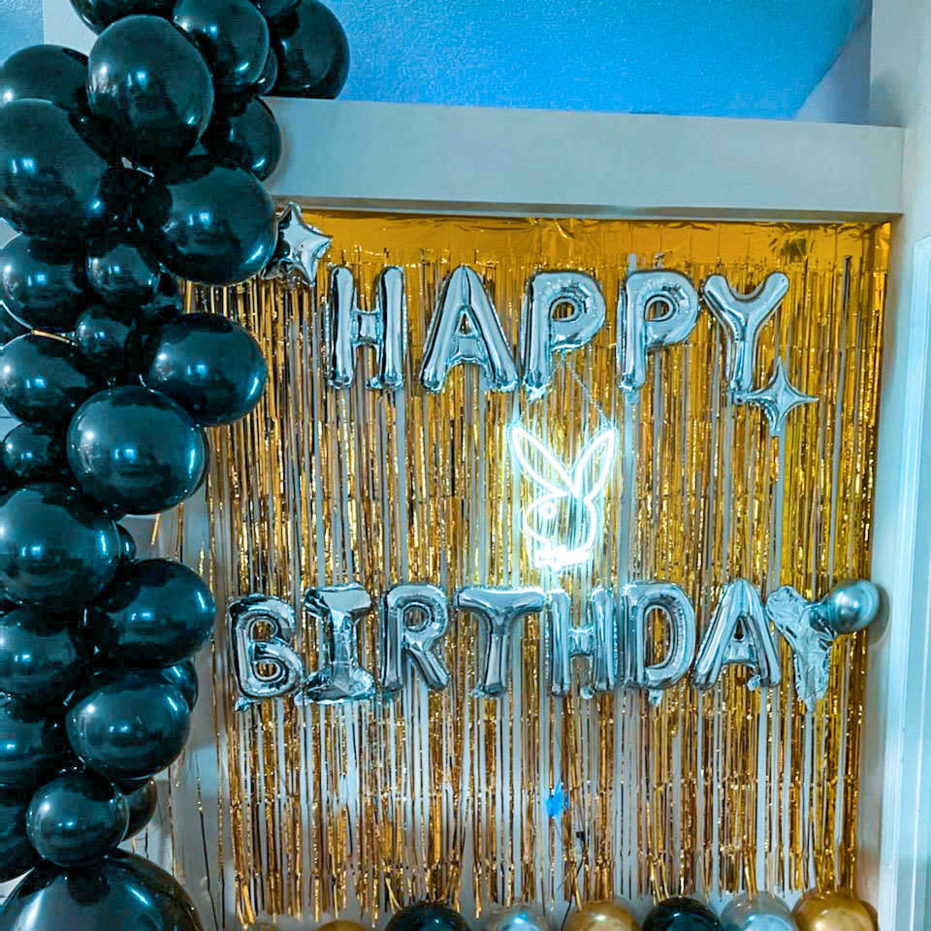 A birthday decoration setup with metallic balloons in black, gold, and silver, alongside a shiny gold fringe backdrop and silver letters spelling "HAPPY BIRTHDAY."