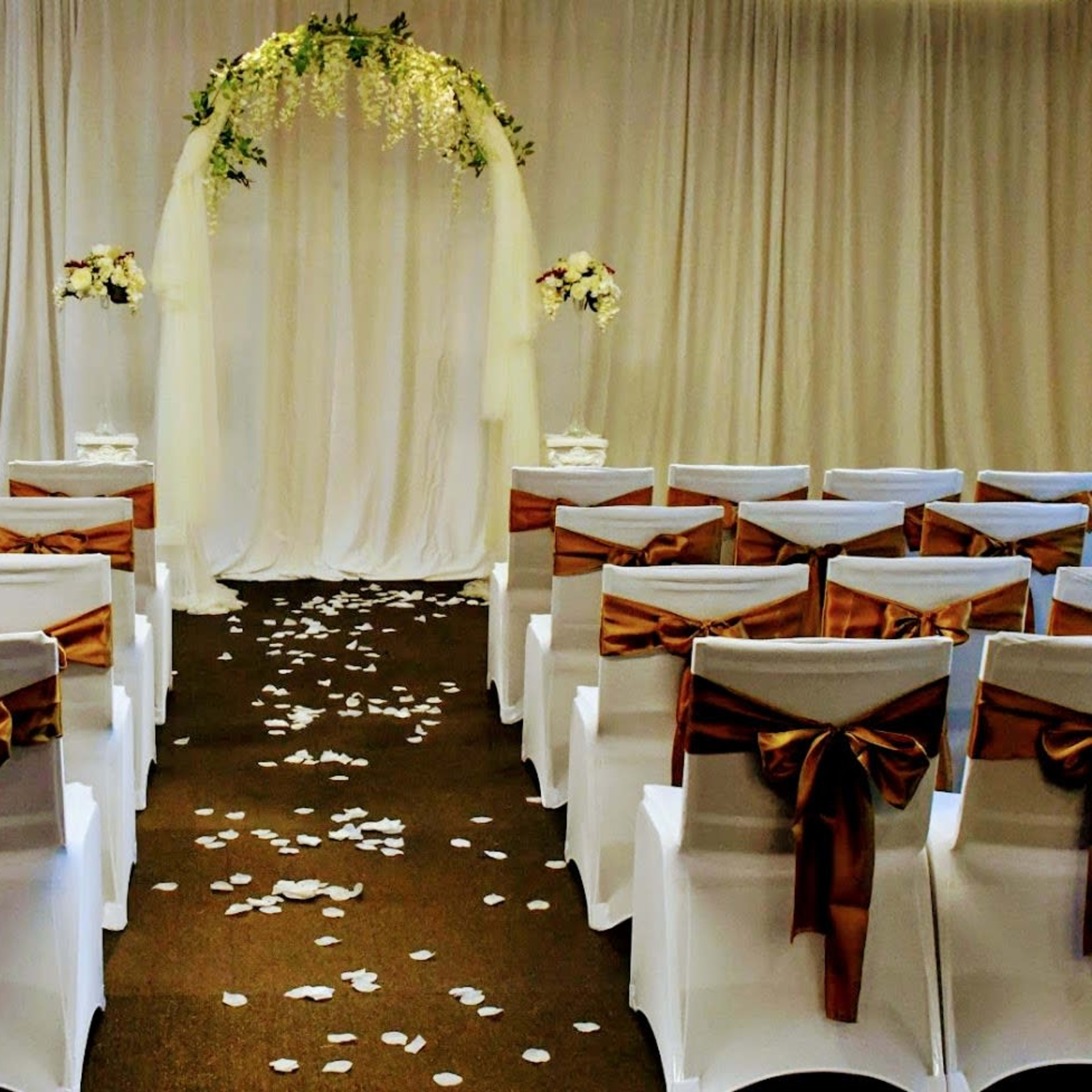 A wedding ceremony setup with white chairs adorned with brown sashes, facing an arch decorated with greenery and flowers, set against a draped backdrop.