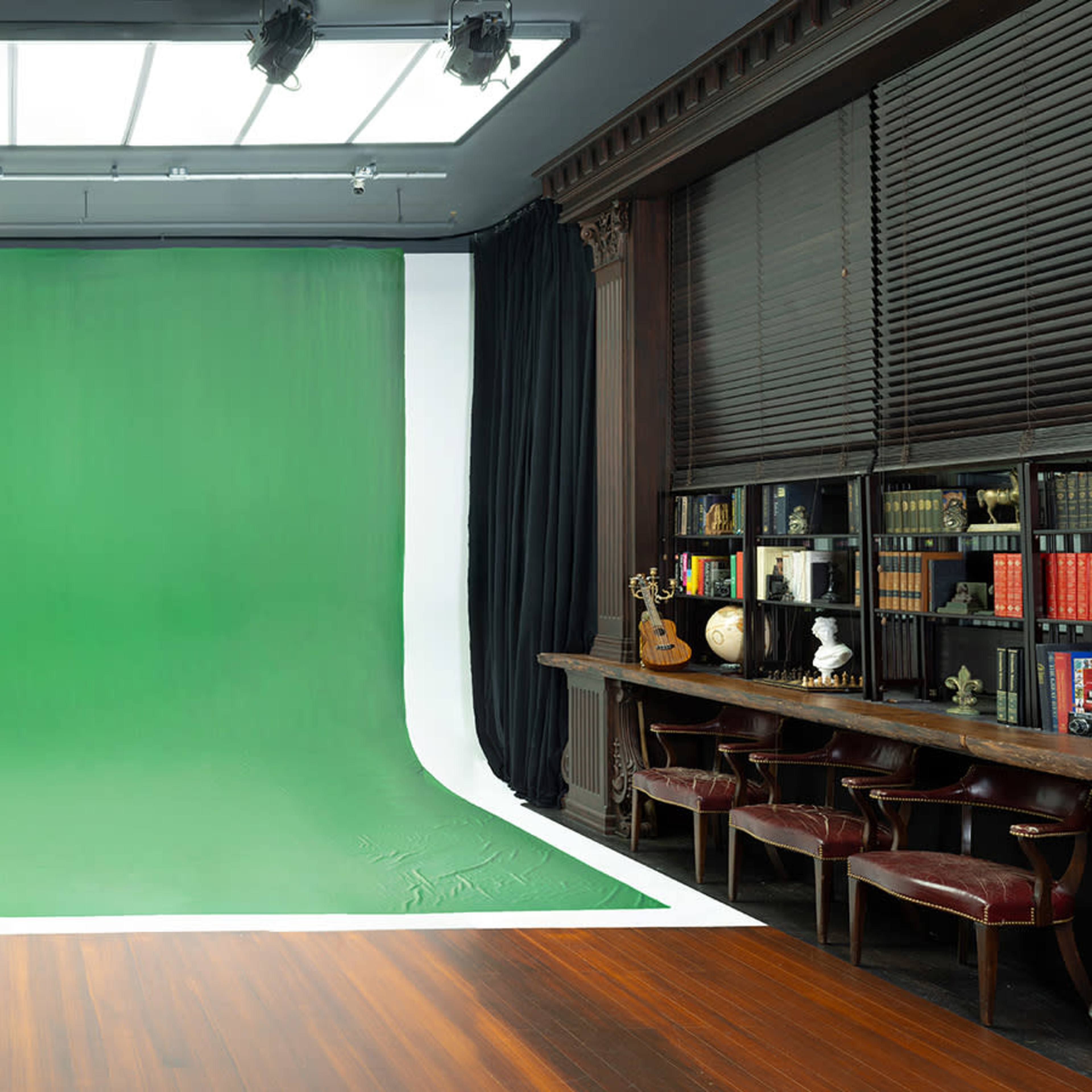 A studio space featuring a large green backdrop, wooden flooring, and a long table lined with books and decorative items.