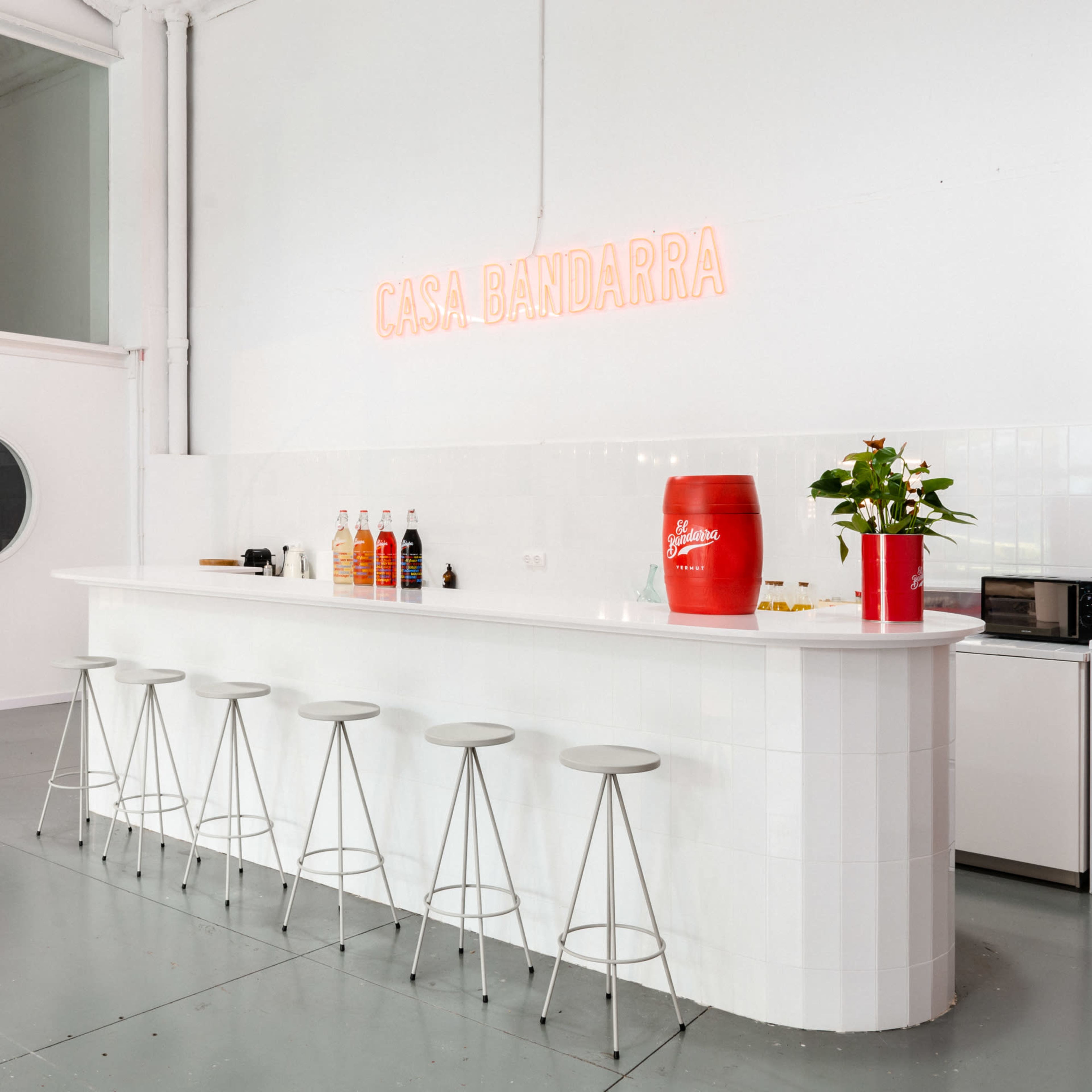 A minimalist white kitchen features a long bar with six stools, a red vase with flowers, and a neon sign reading "CASA BANDARRA."