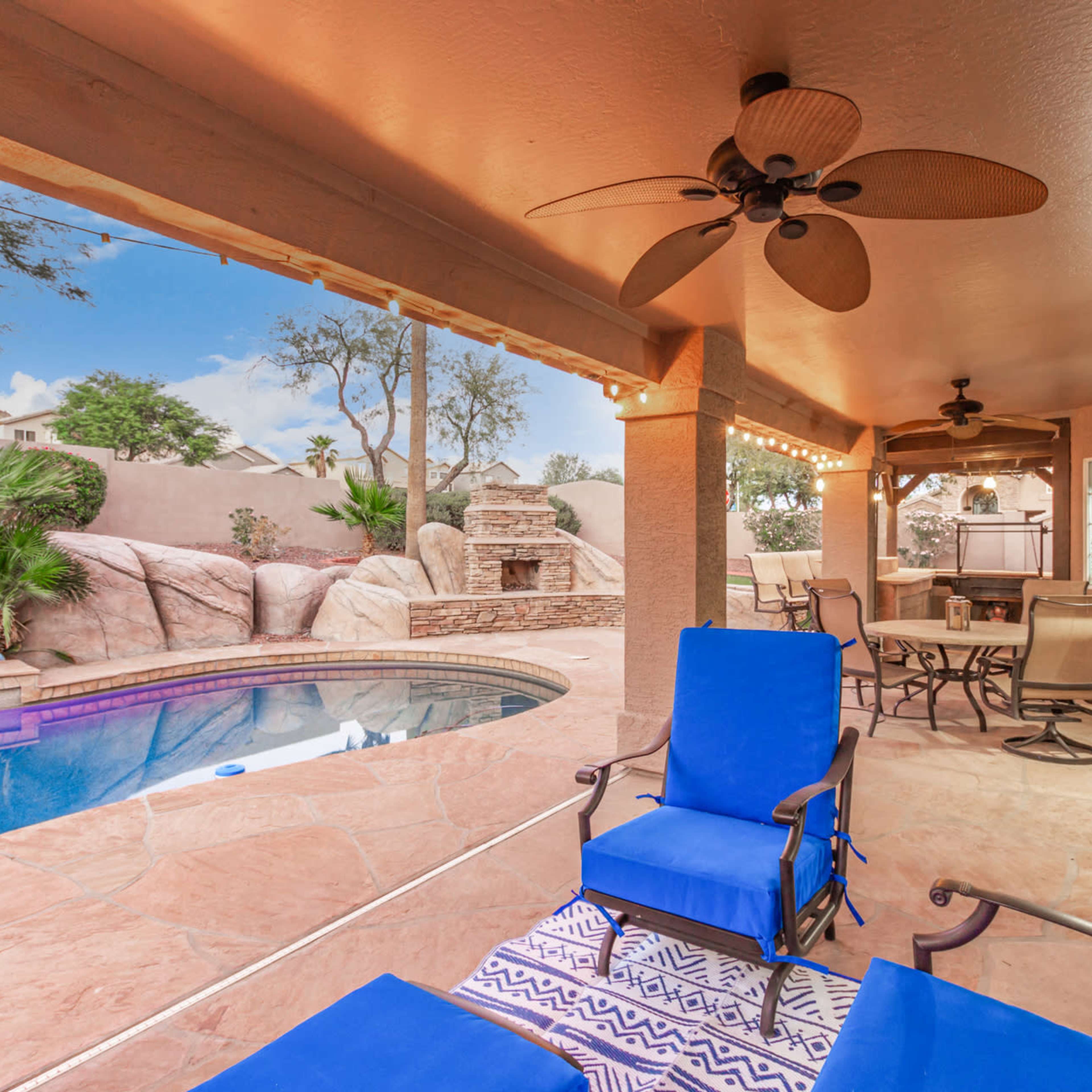 A covered patio with blue lounge chairs overlooks a swimming pool and landscaped yard featuring rock formations and a stone grill area.
