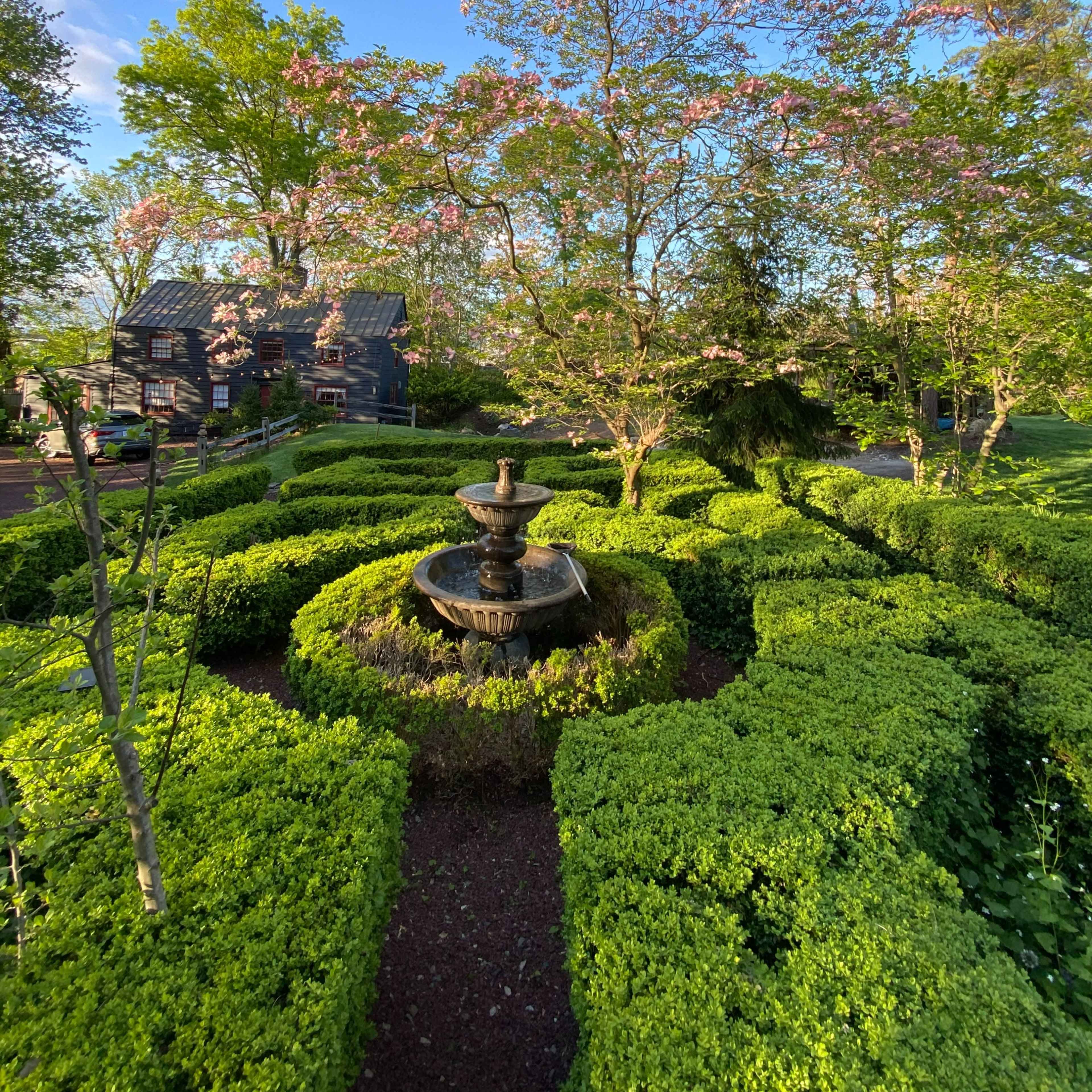 A neatly manicured garden features a central fountain surrounded by trimmed hedges and blooming trees.
