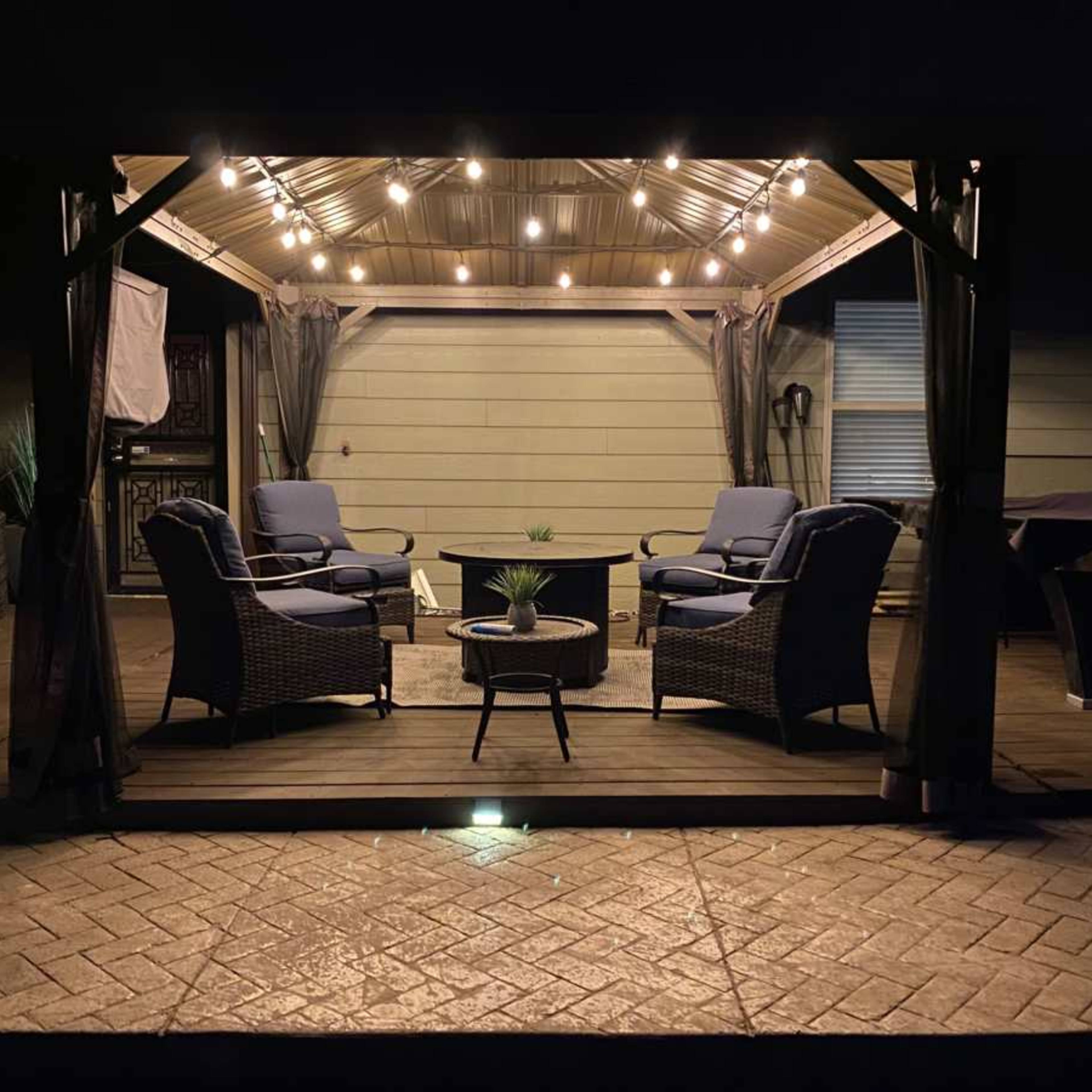 A covered outdoor seating area features two chairs and a table, illuminated by string lights against a twilight sky.