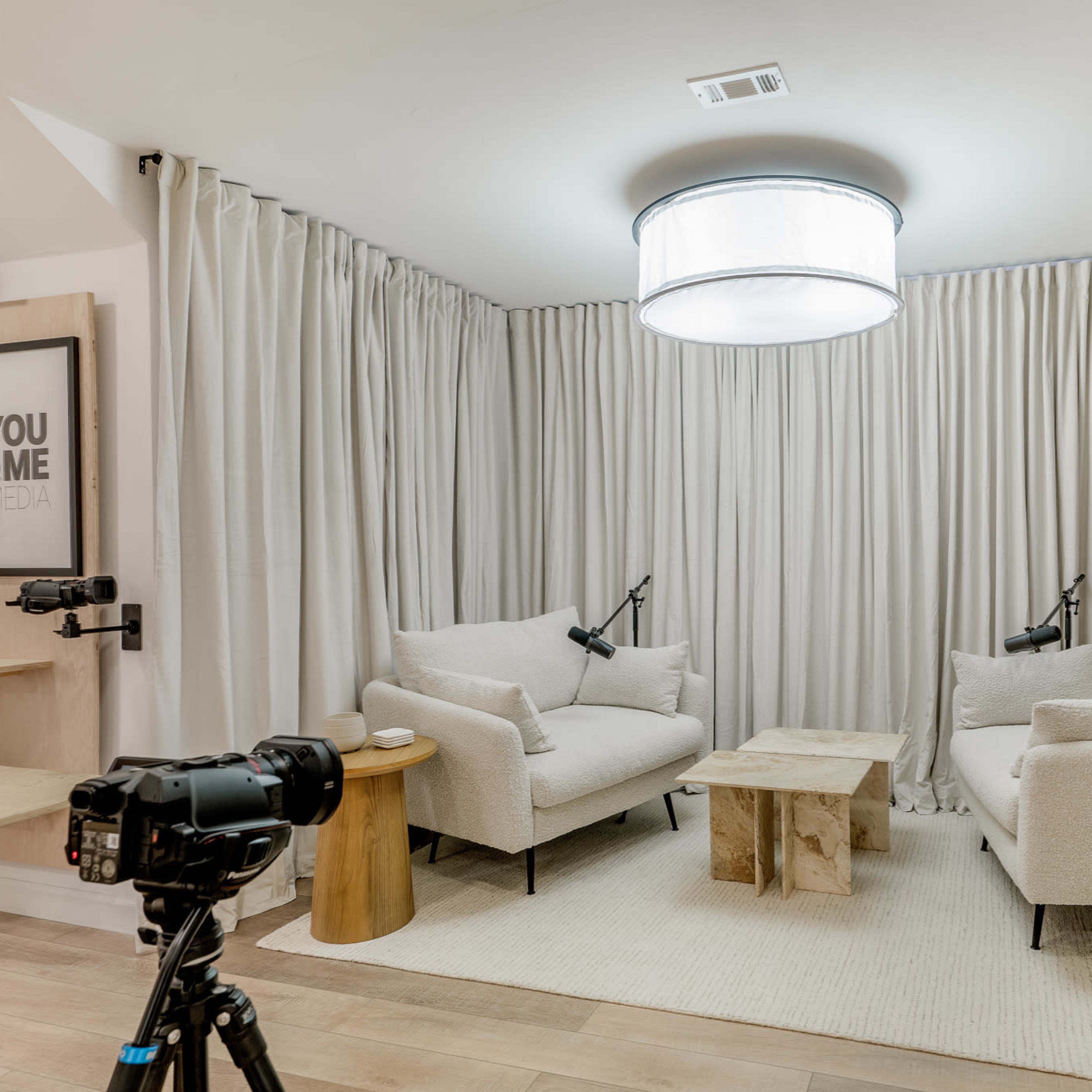 A seating area with two light-colored armchairs facing a wooden coffee table, surrounded by beige curtains and equipped with two mounted cameras.