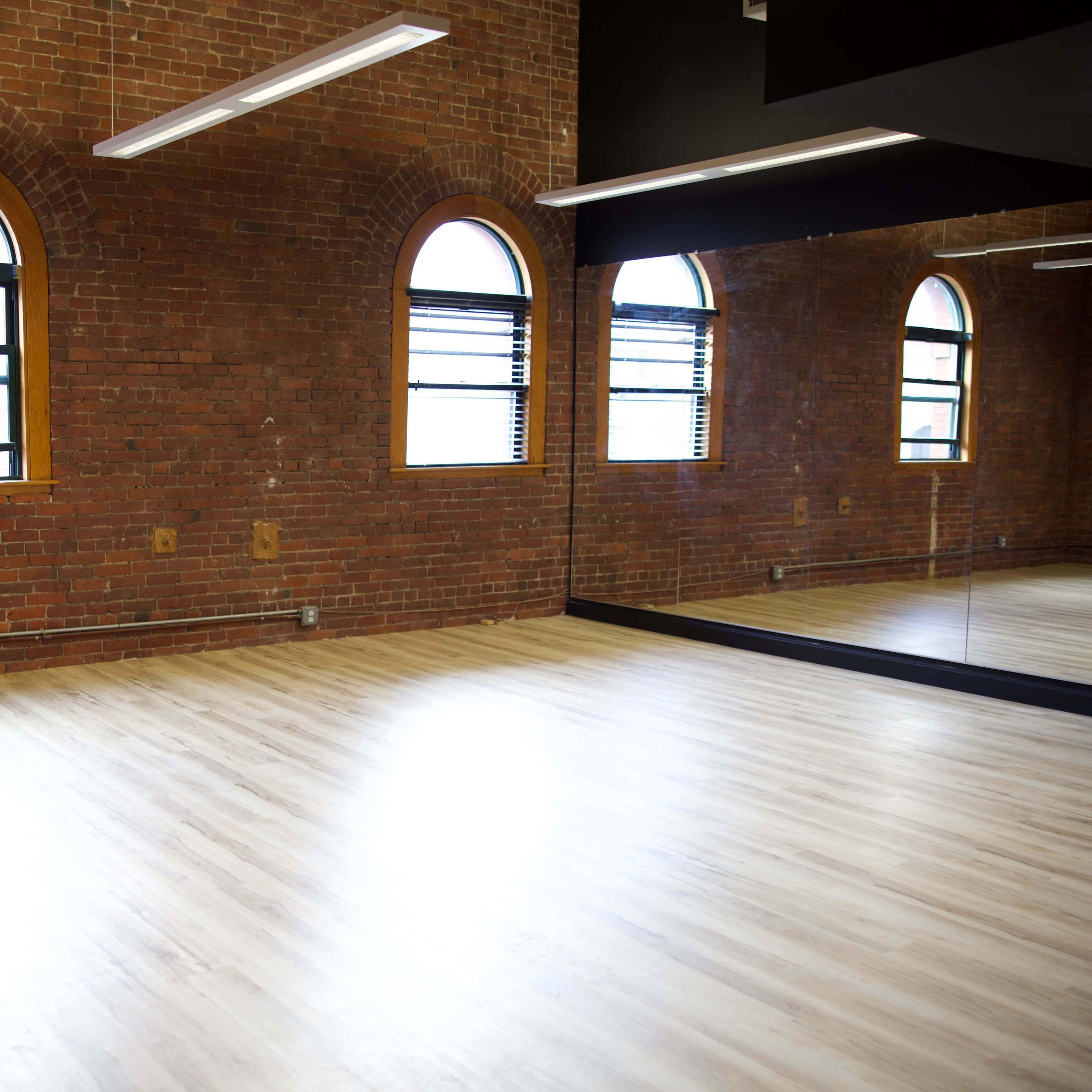 A spacious room with exposed brick walls, large windows, and a mirror along one side.