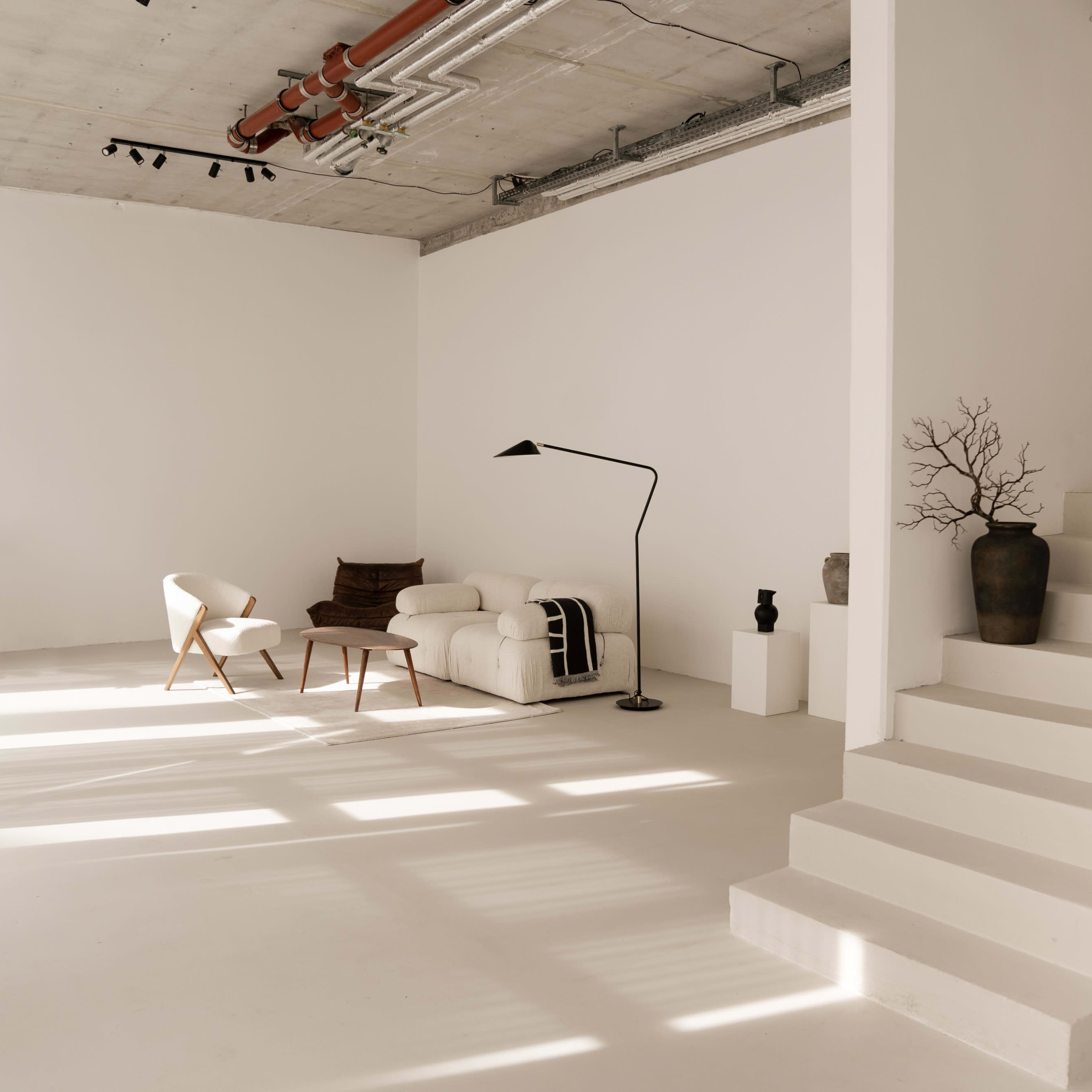 A bright, minimalist room features a white sofa, a chair, a floor lamp, and potted plants beside a staircase.