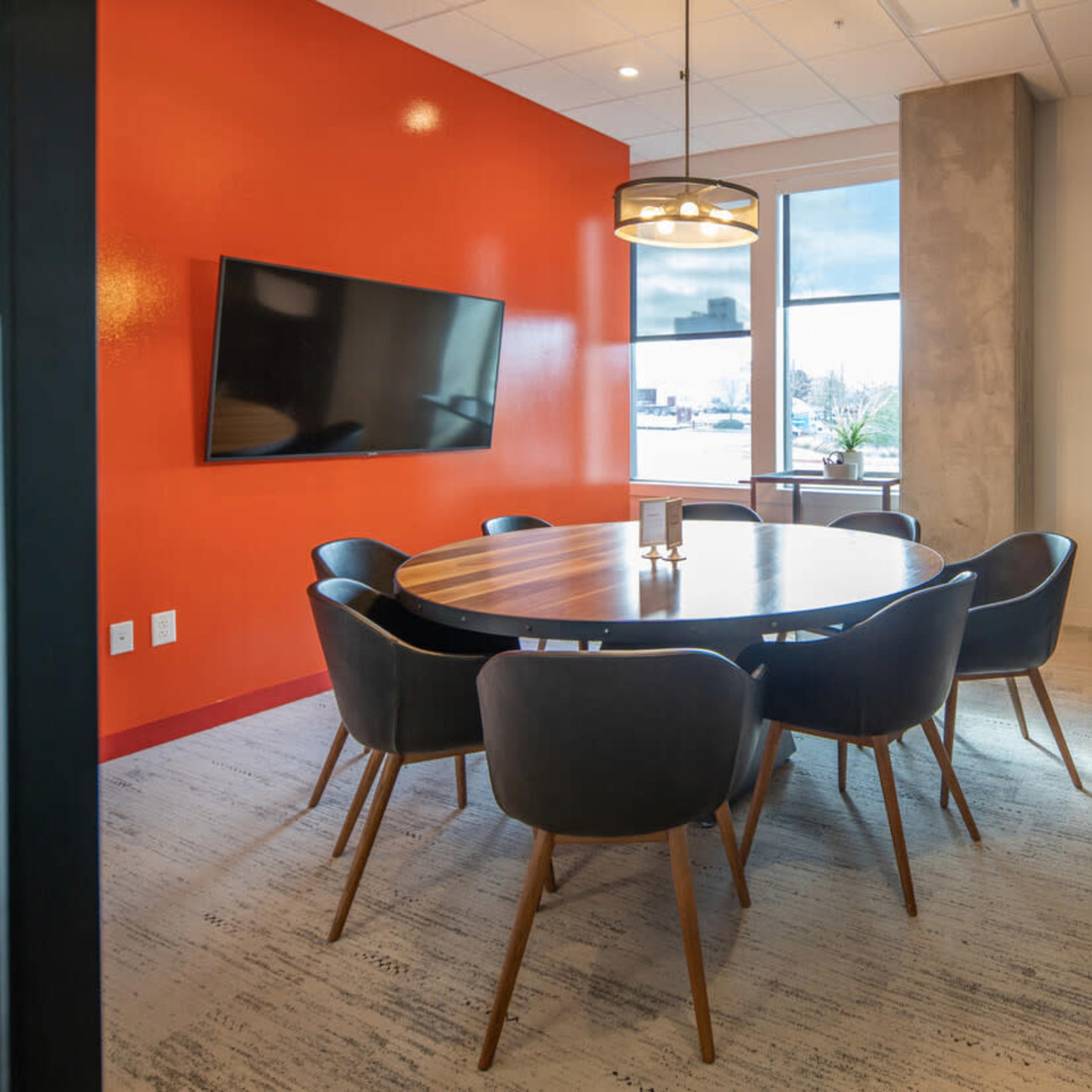 A modern conference room with a round wooden table surrounded by black chairs, an orange accent wall, and a large television mounted on the wall.