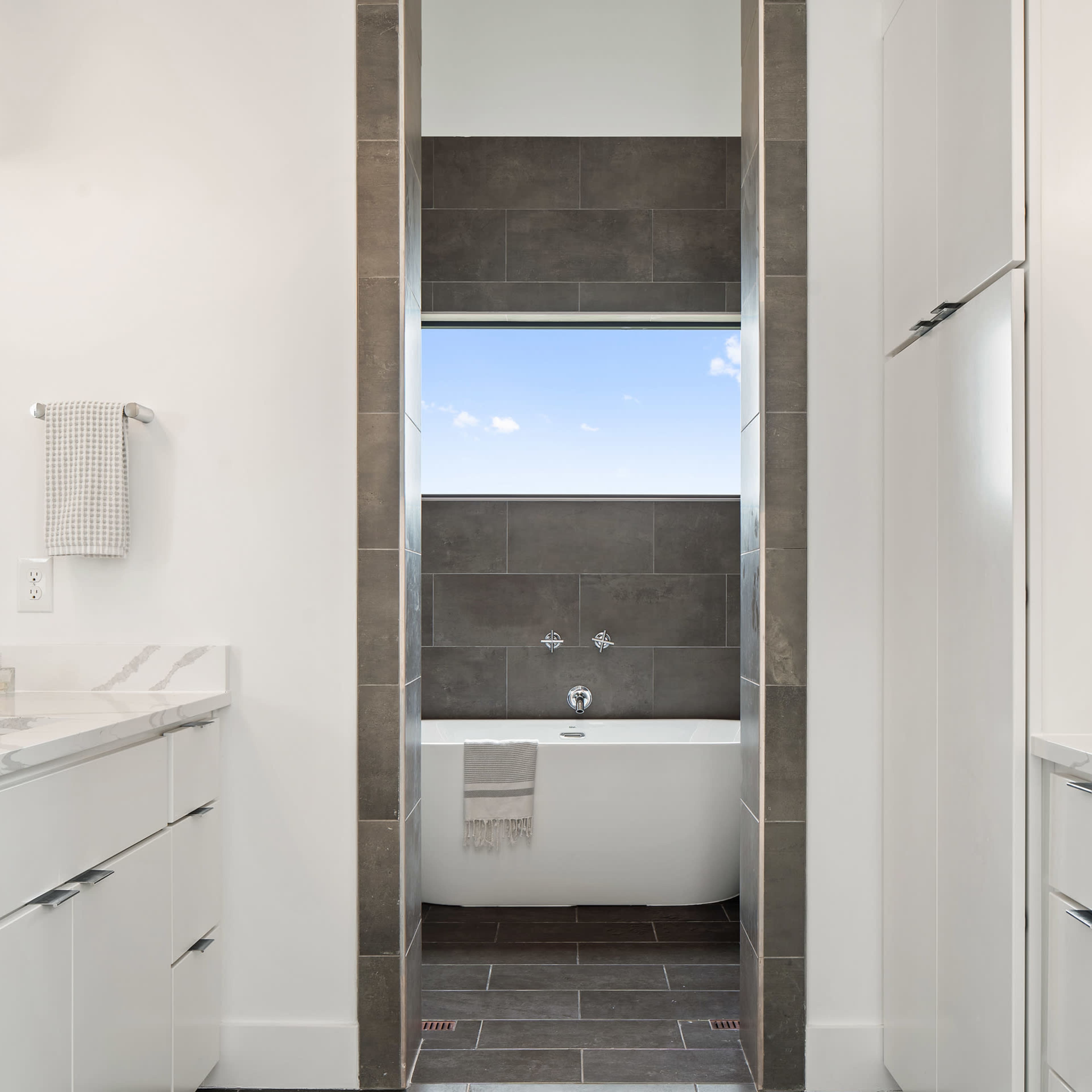 A modern bathroom features a tub surrounded by gray tiles, with white cabinetry on either side and a window above the tub.
