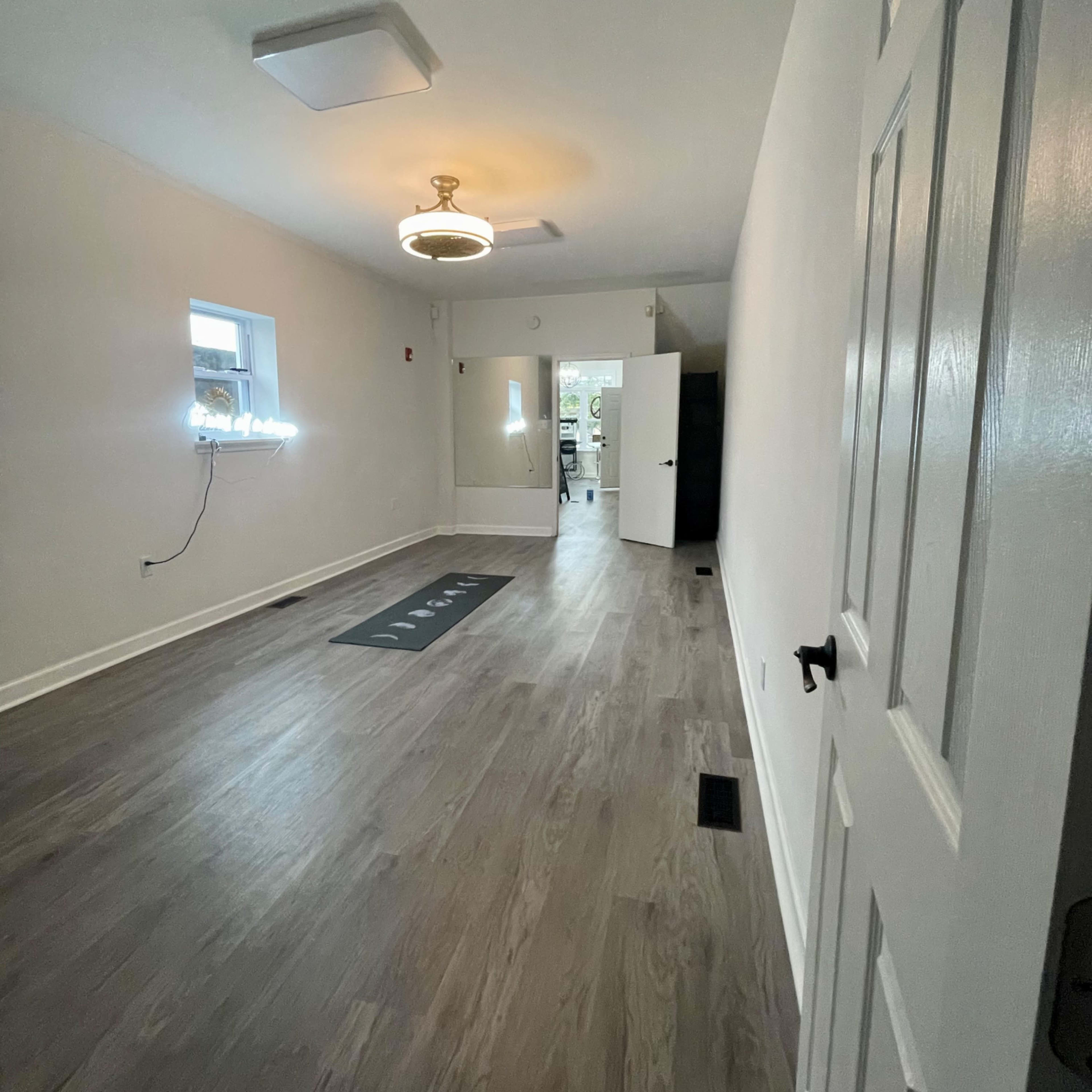 A sparsely furnished room with light-colored walls, a wooden floor, a ceiling light, and a doorway leading to another space.