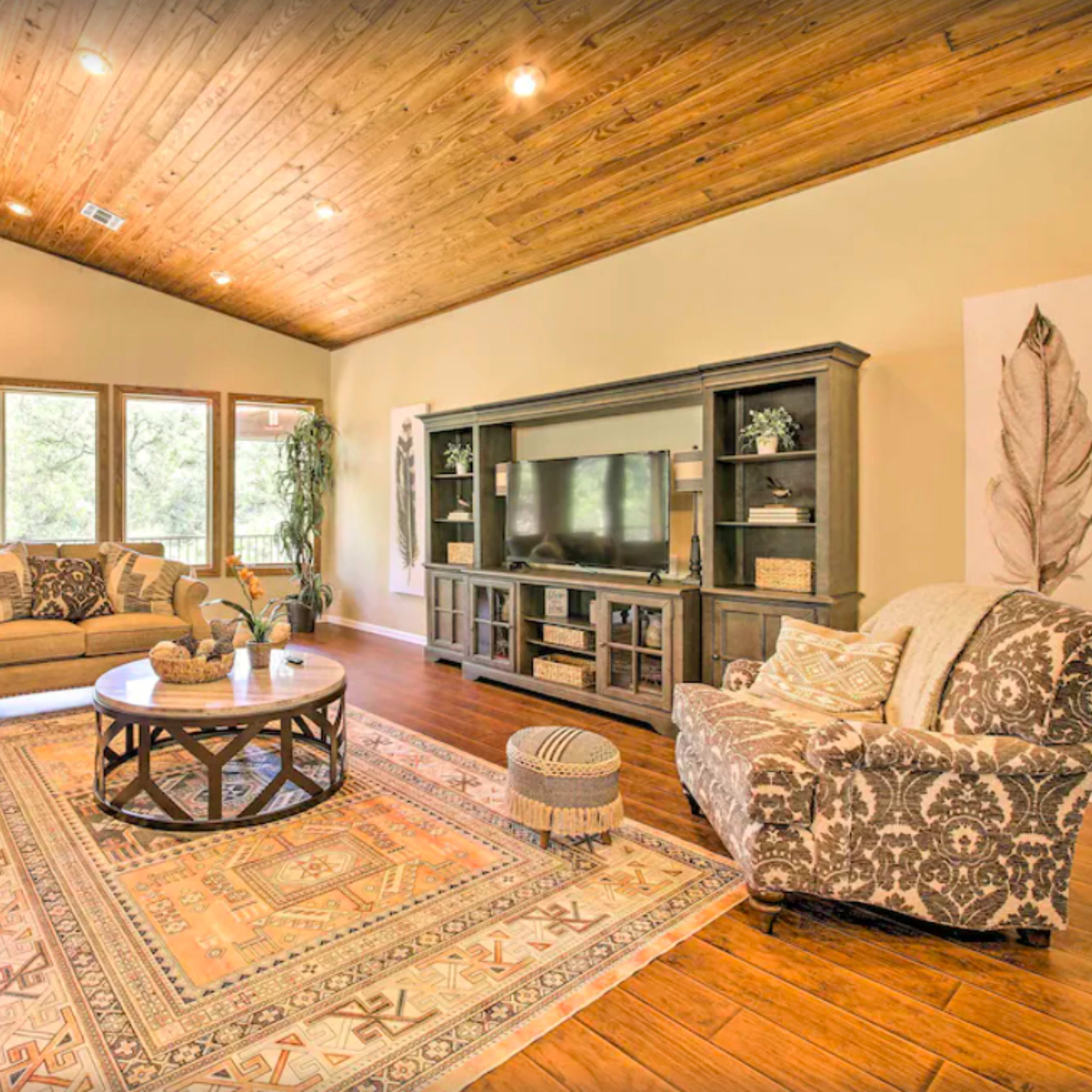 The living room features a large area rug, a circular coffee table, and a media console against a wall of windows that offer a view of the outdoors.