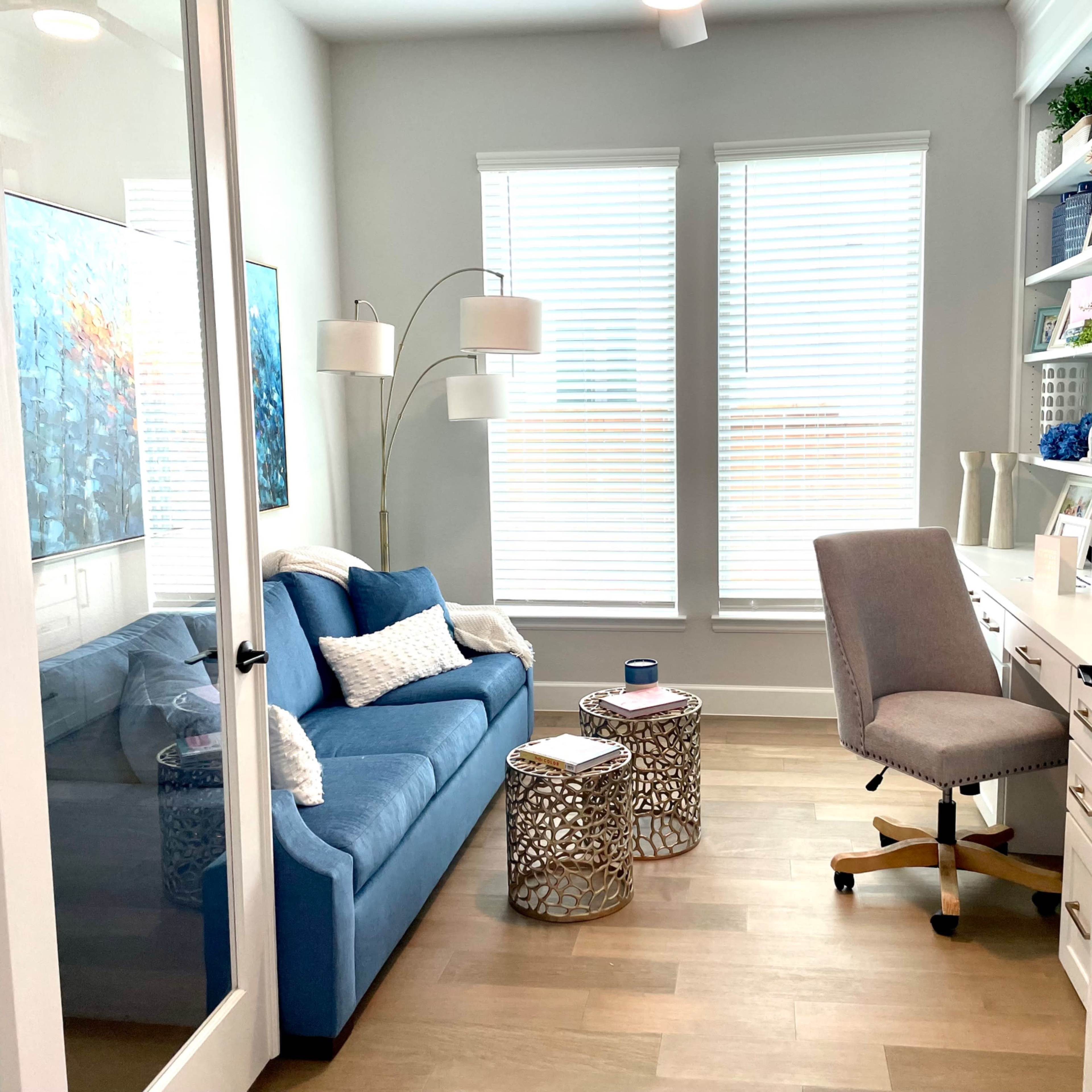 A small office space features a blue couch, two decorative tables, a desk with a chair, and large windows with white blinds.