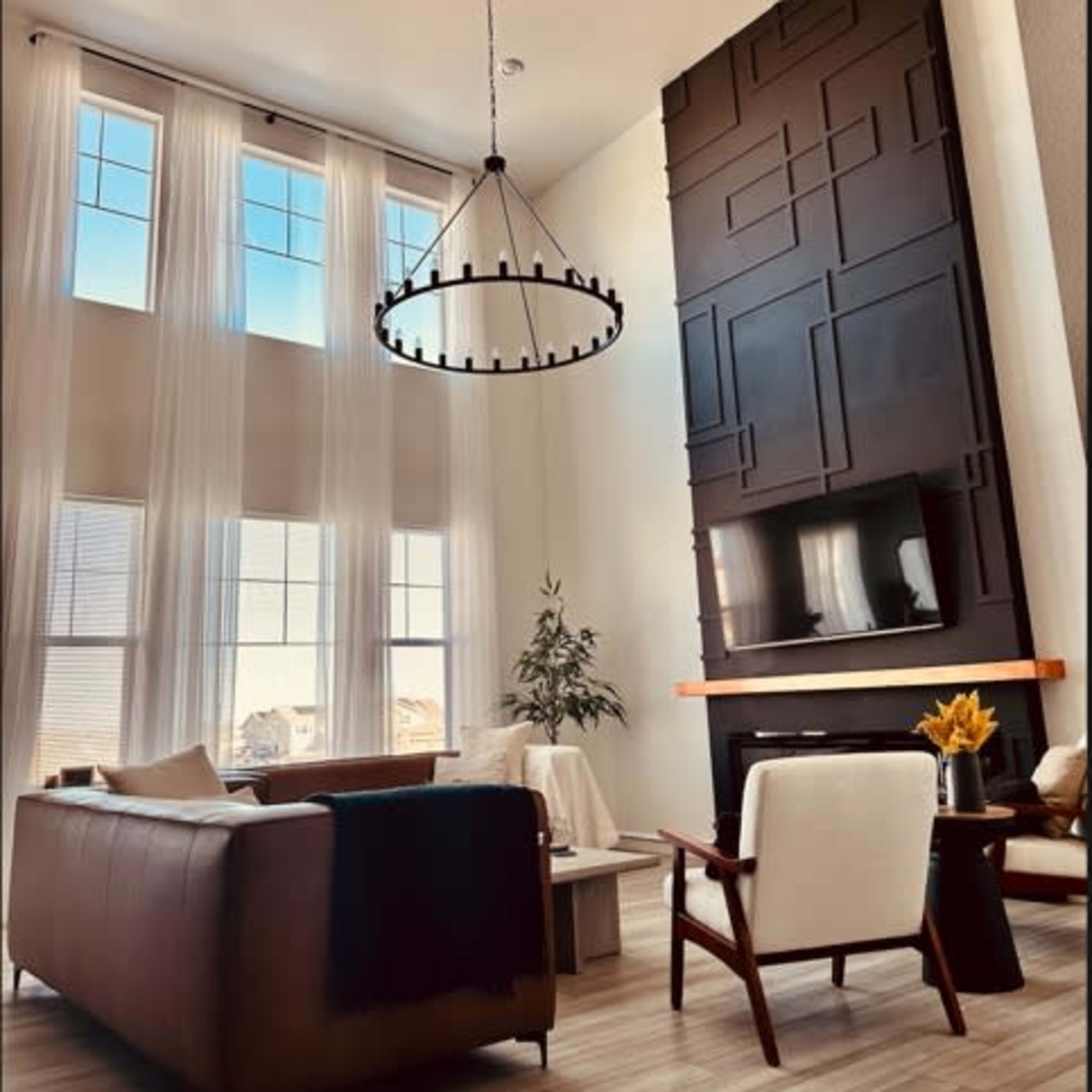 A modern living room with high ceilings, large windows, a circular chandelier, and a dark accent wall with a fireplace.
