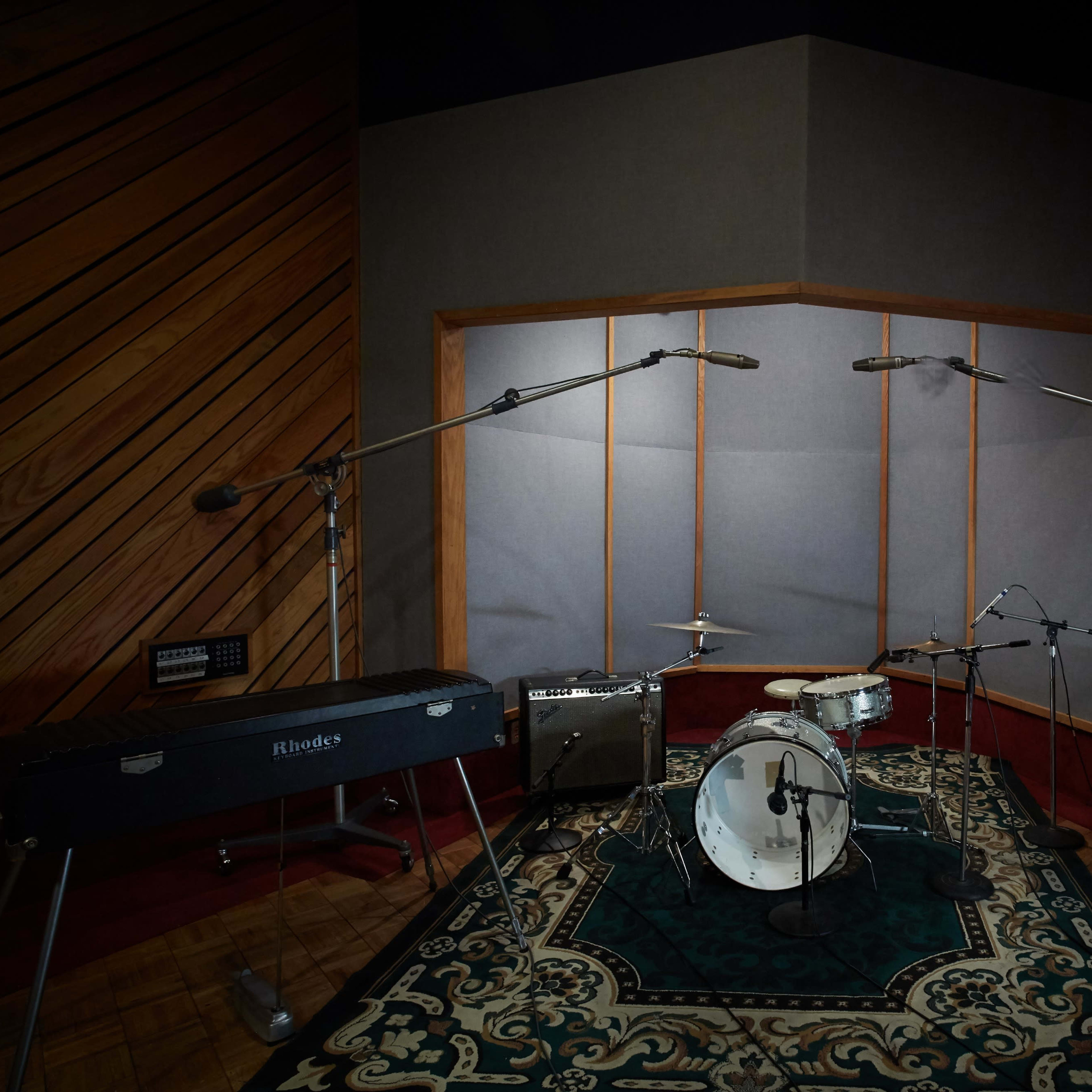 A recording studio with a drum set, microphones, amplifiers, and a keyboard on a patterned rug.