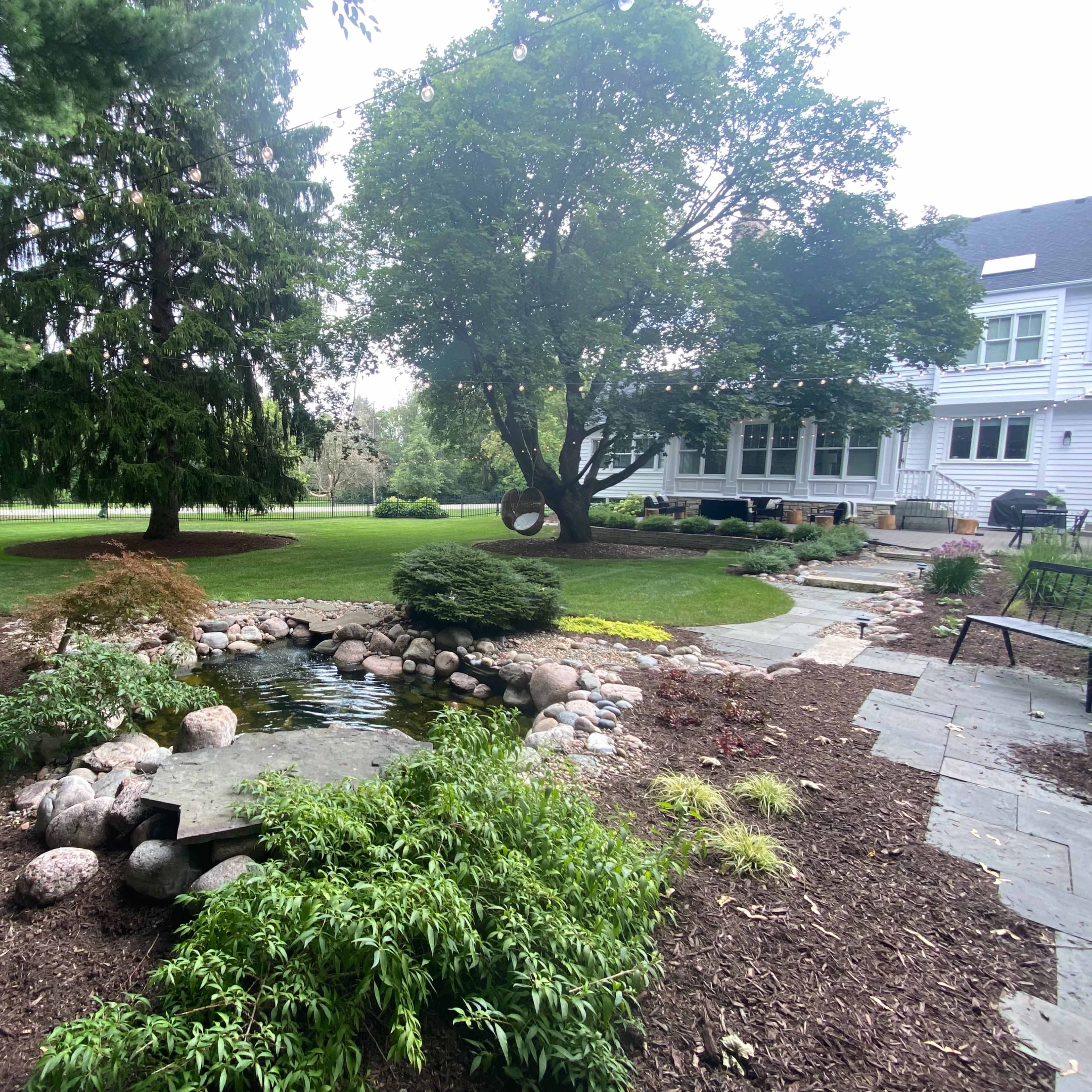A landscaped backyard featuring a small pond surrounded by rocks, a grassy area, and various trees and shrubs.