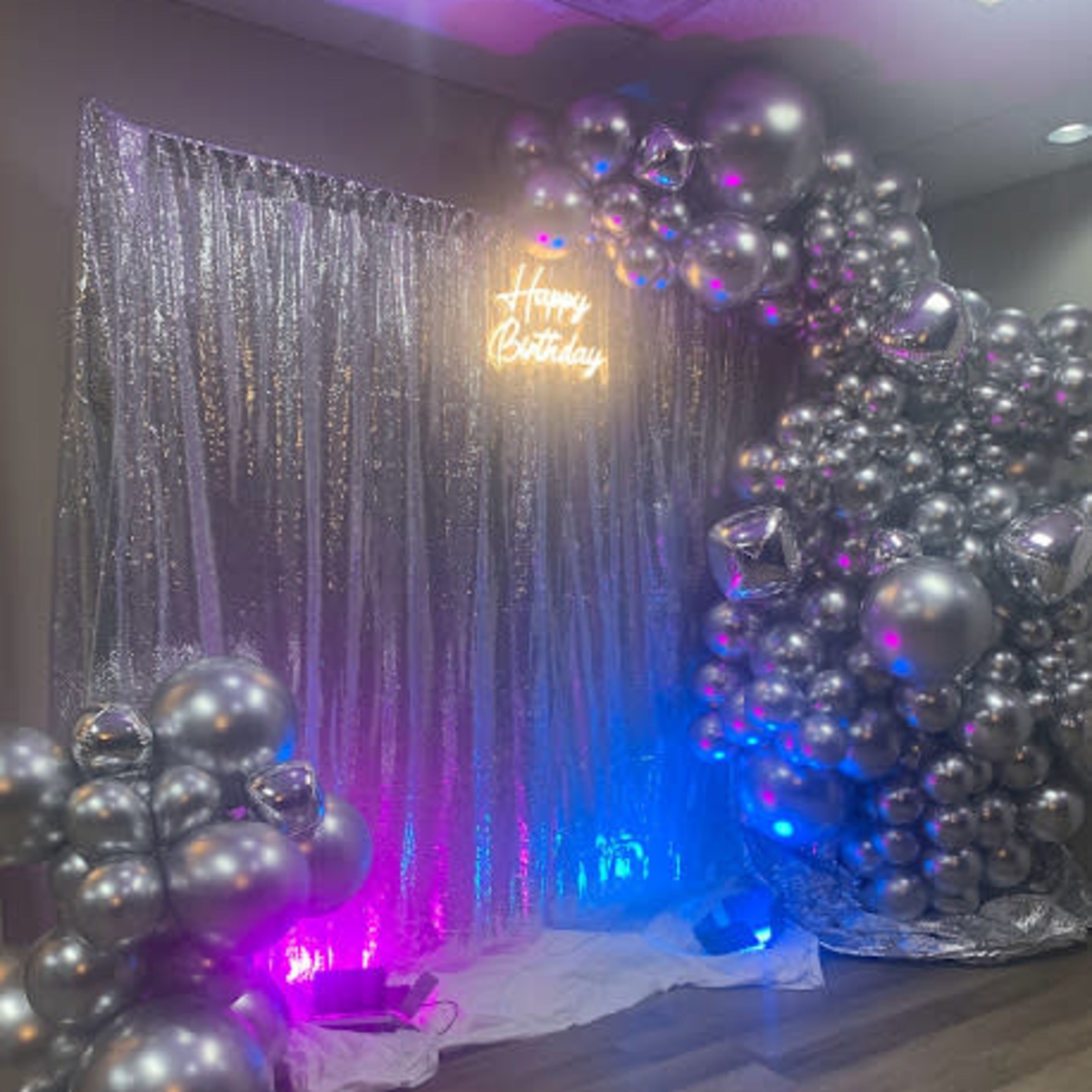 A decorative backdrop for a birthday celebration, featuring a silver curtain, neon "Happy Birthday" sign, and an arrangement of metallic balloons.