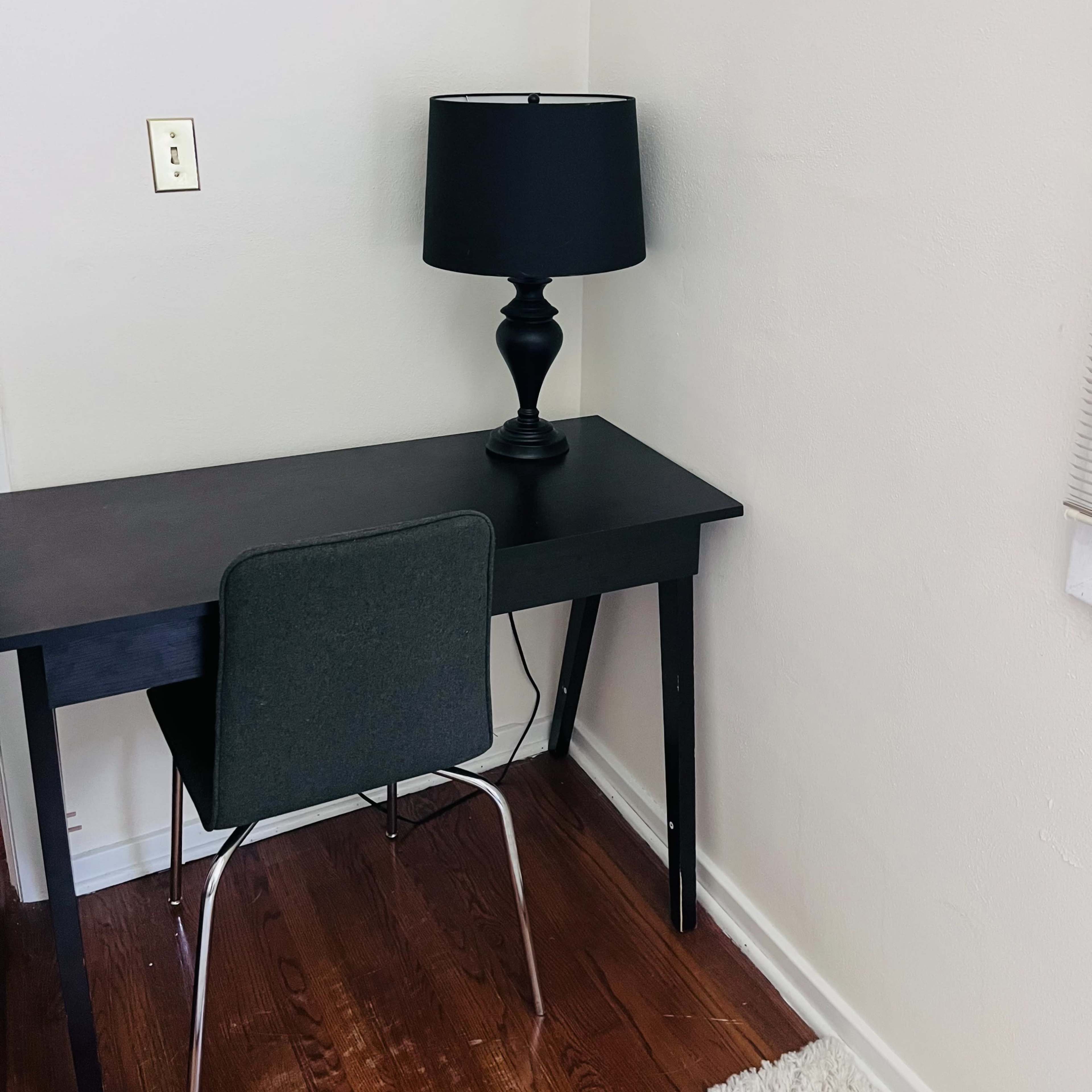A black desk with a matching lamp and a gray chair is positioned in a corner of a room with light-colored walls and hardwood flooring.