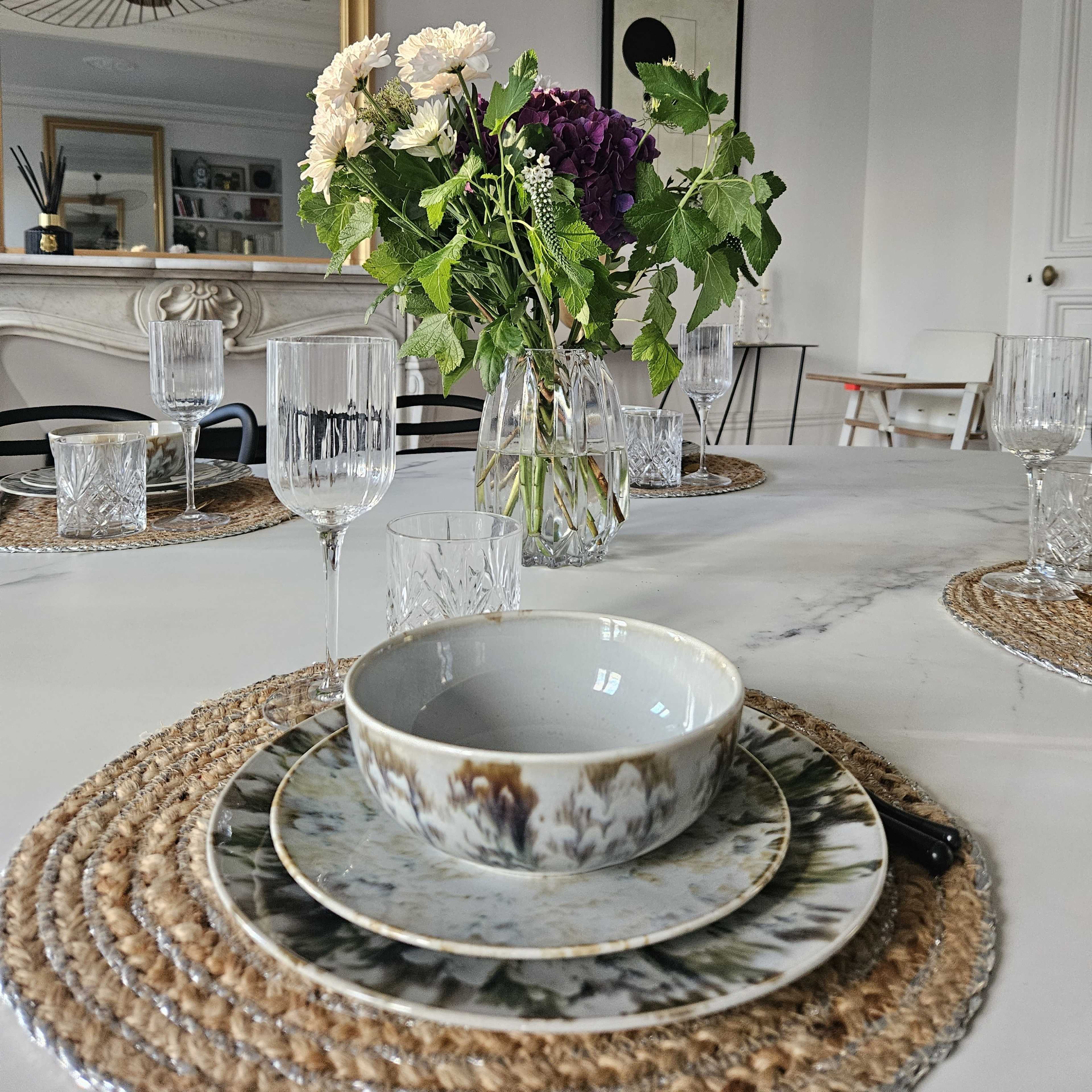 A dining table is set with a decorative plate and bowl, surrounded by crystal glasses and a vase of flowers, all placed on a woven placemat.