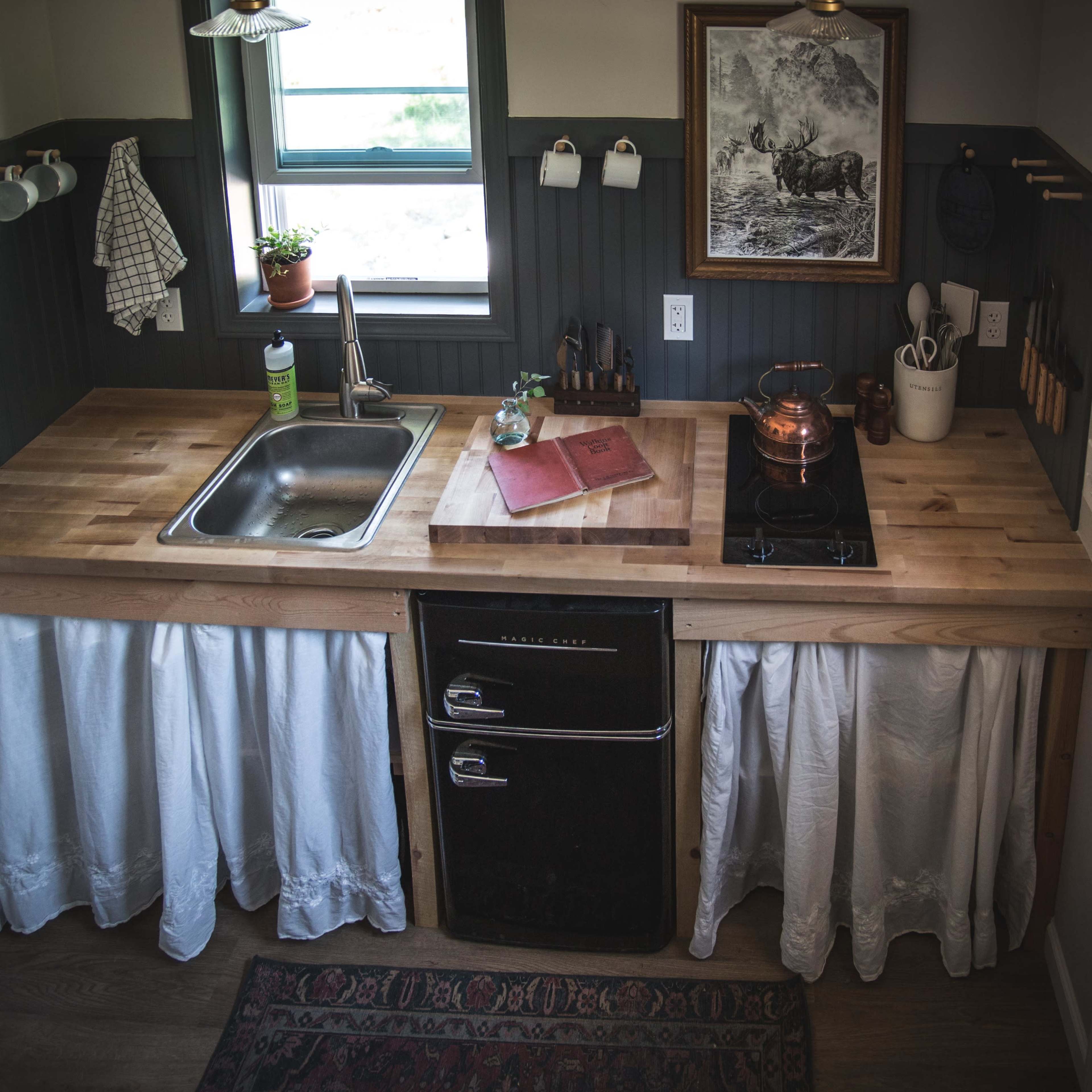 A compact kitchen with a wooden countertop, a sink, a stovetop, and a refrigerator beneath a white curtain, set against dark green wall panels and framed by a door on the right.