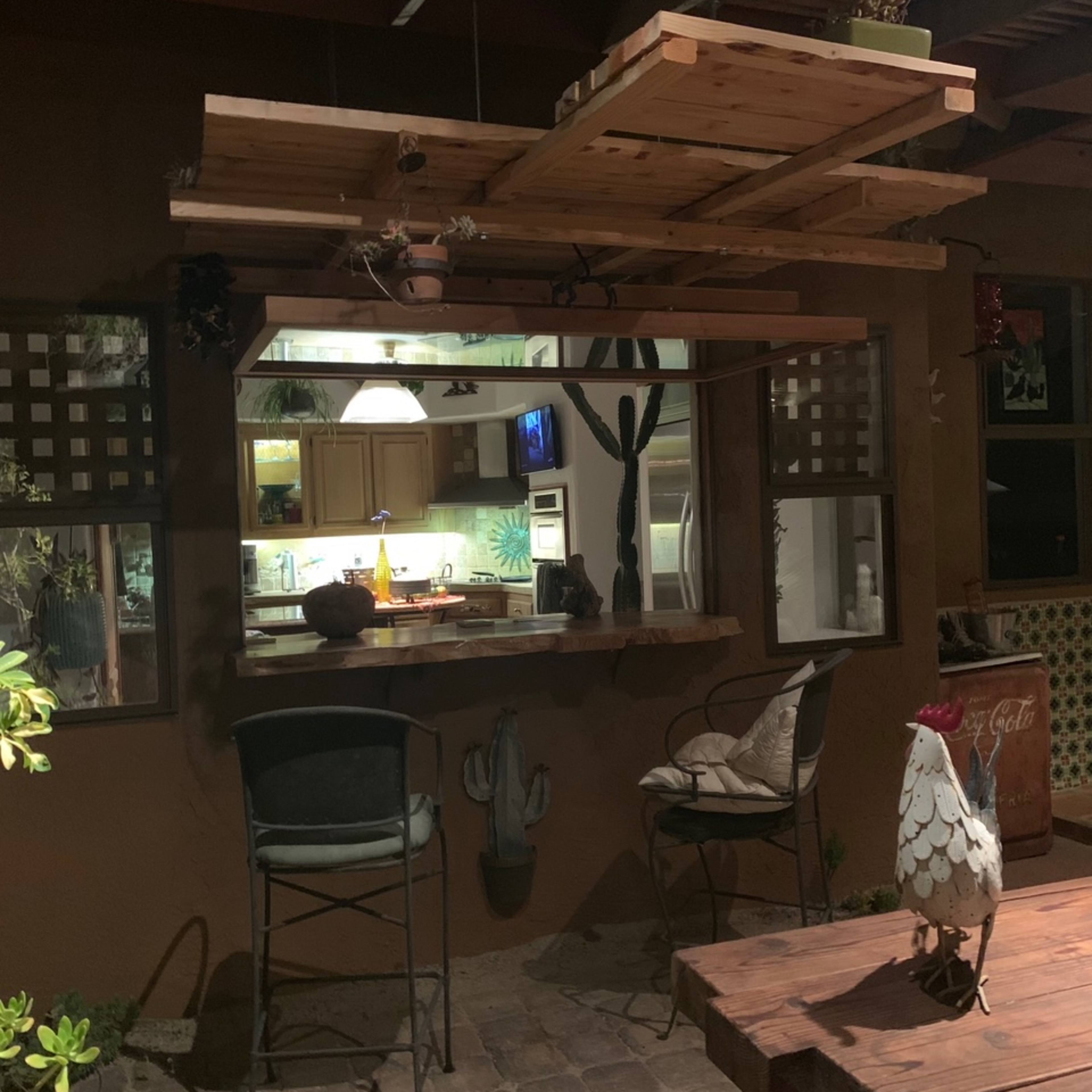 An outdoor bar area with a wooden countertop, high stools, and a view into a kitchen illuminated by warm light.