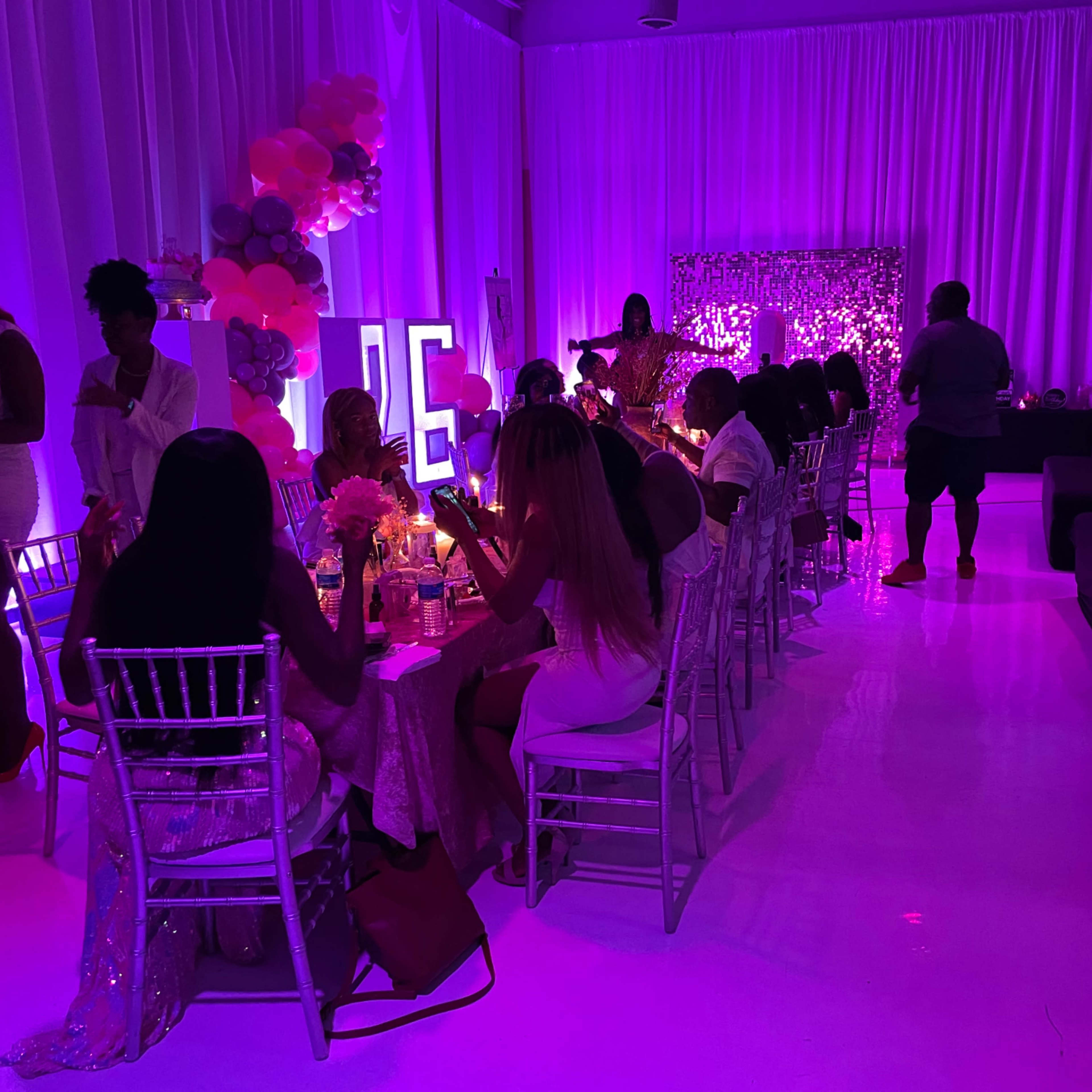 A group of people sit at a long table in a brightly lit event space decorated with pink balloons and a backdrop.
