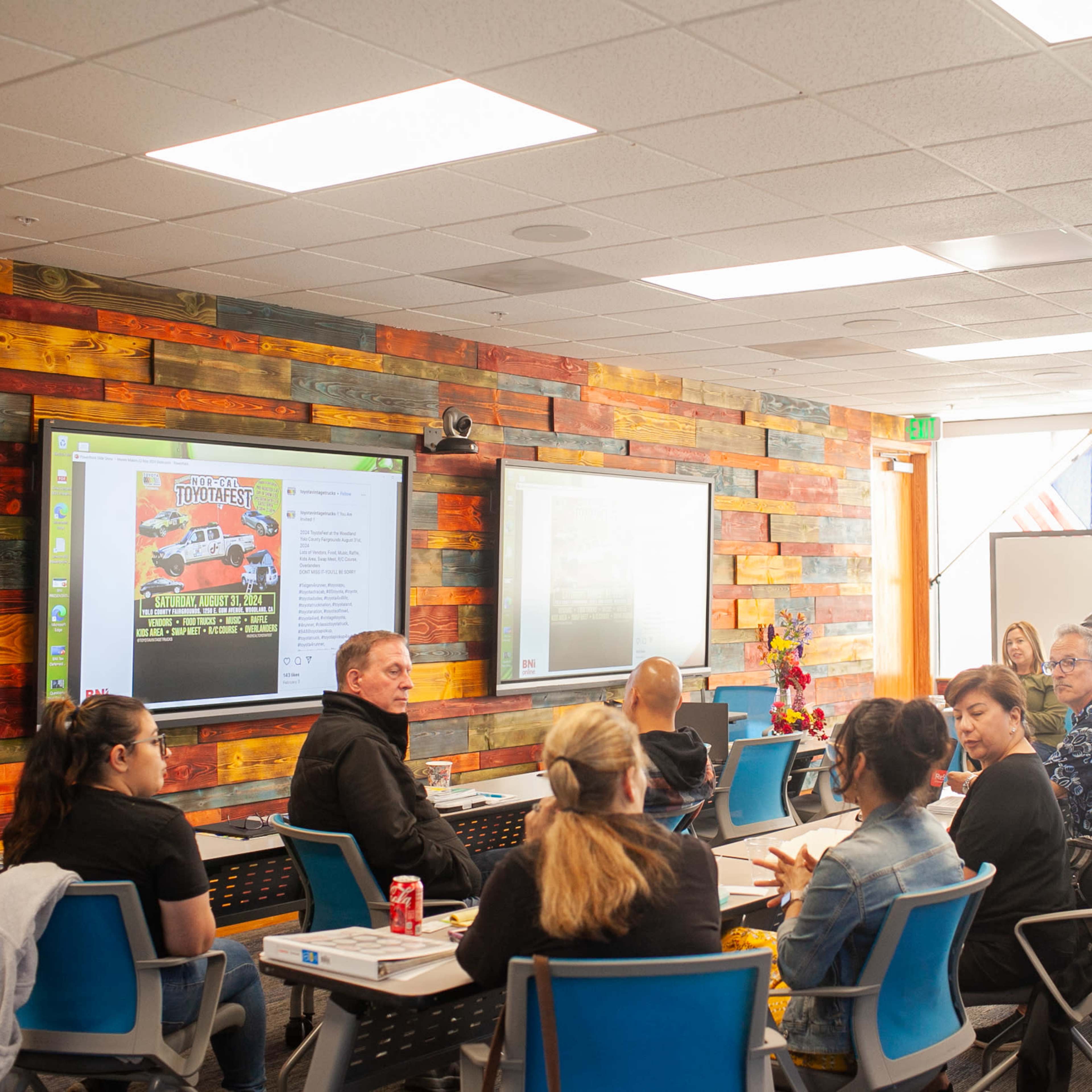 A group of people is seated in a classroom with wooden-paneled walls, engaging in discussion while facing a screen displaying promotional material.