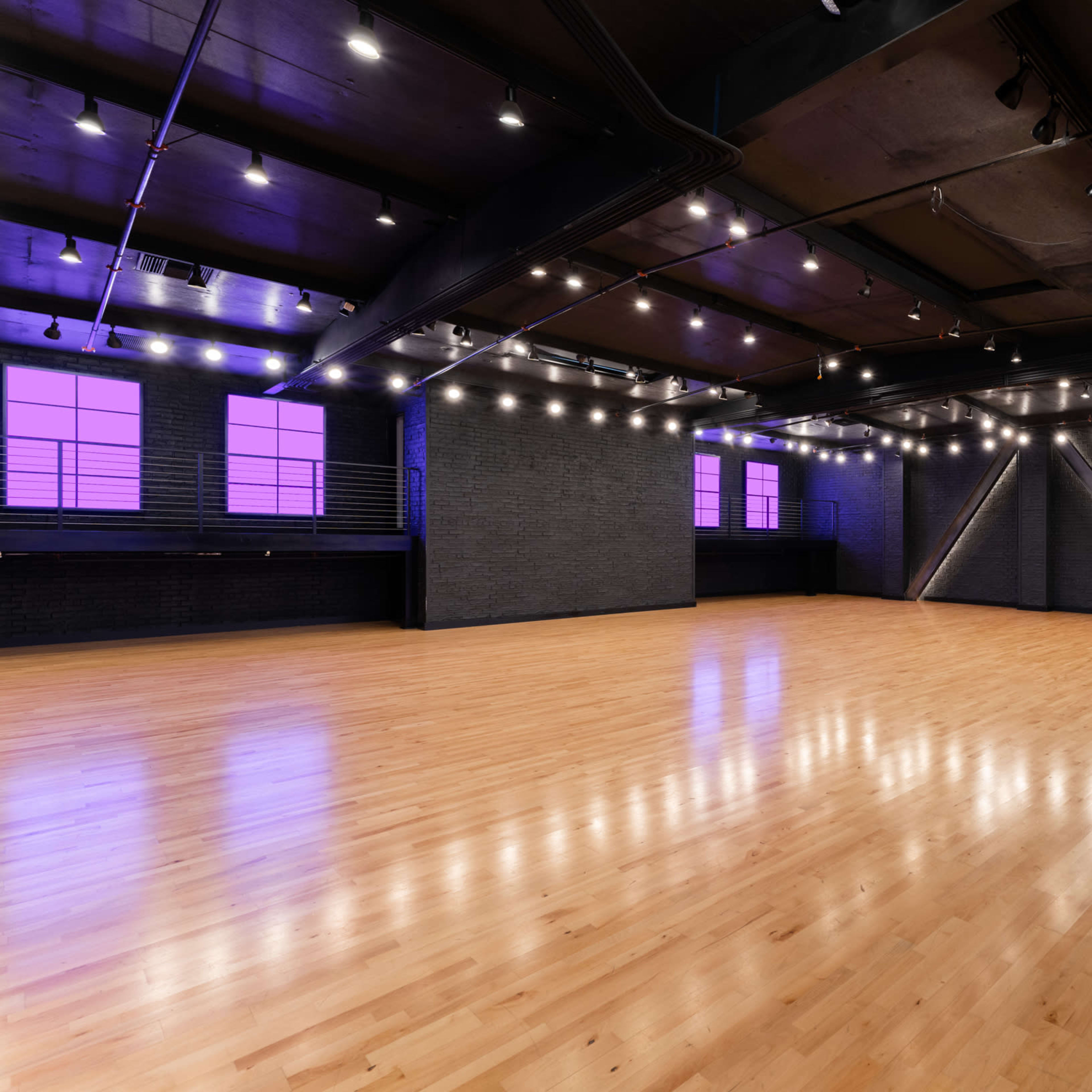 A spacious dance studio with polished wooden floors, black walls, and large purple windows, along with two pink giraffe sculptures in the corner.