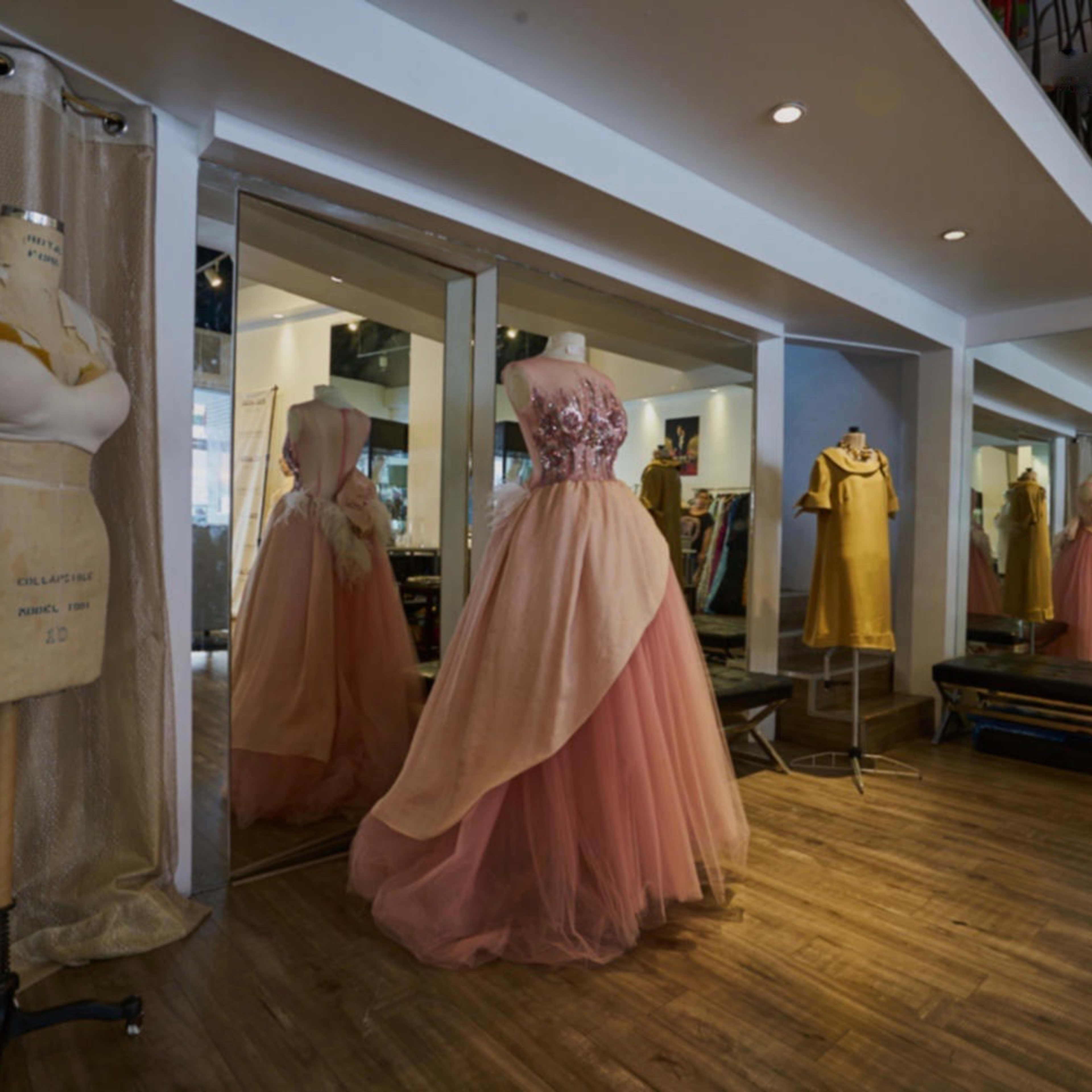 A fashion boutique interior featuring a collection of dresses displayed on mannequins and hangers, with mirrors and wooden flooring.