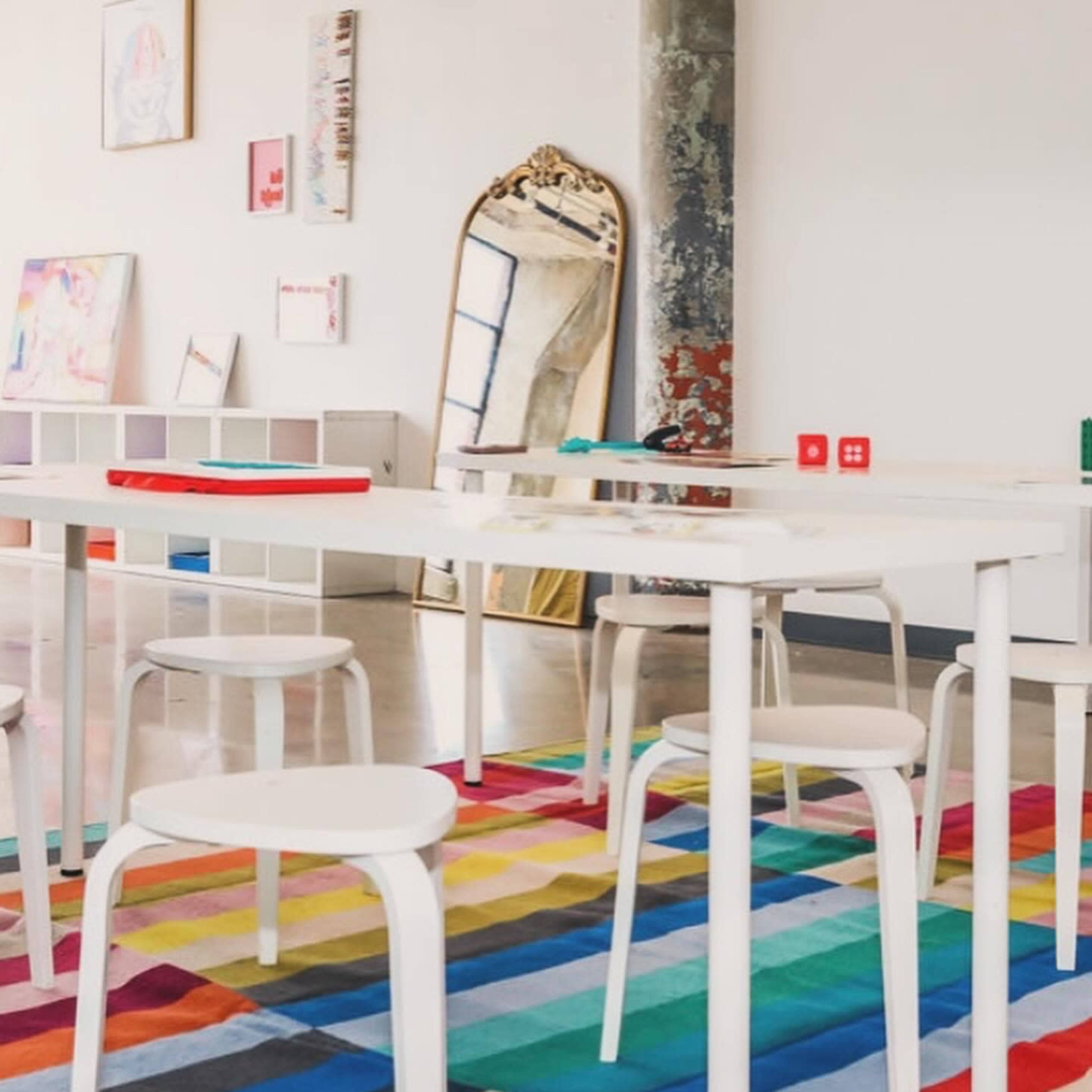 The scene features a brightly colored, patterned rug on a polished floor, with white tables and stools arranged around a large ornate mirror against a light-colored wall.