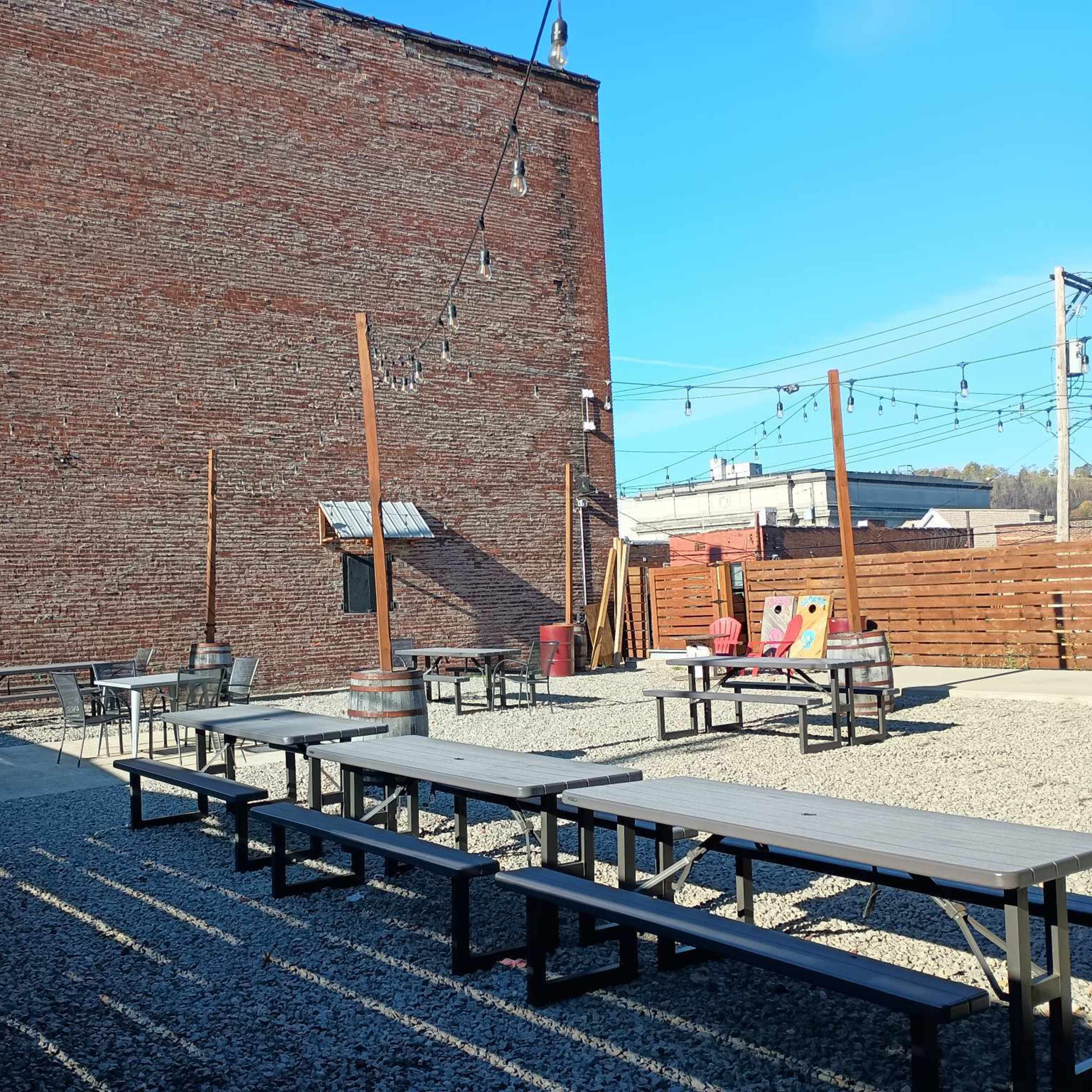 An outdoor seating area with several picnic tables and benches set on gravel, against a brick wall and under clear blue skies.