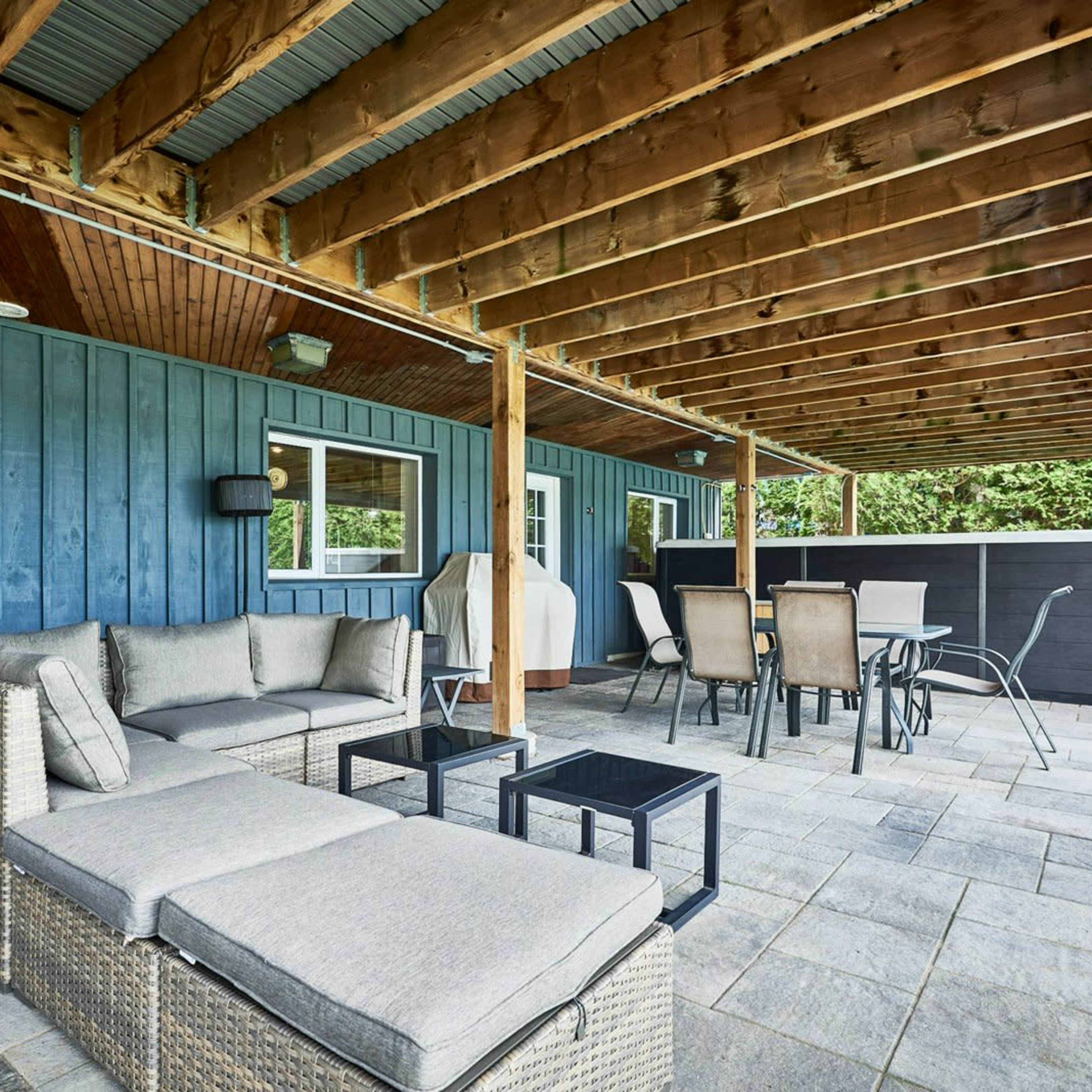 A covered patio area featuring a sectional sofa, a glass table, and dining chairs arranged on a stone surface beneath wooden beams.
