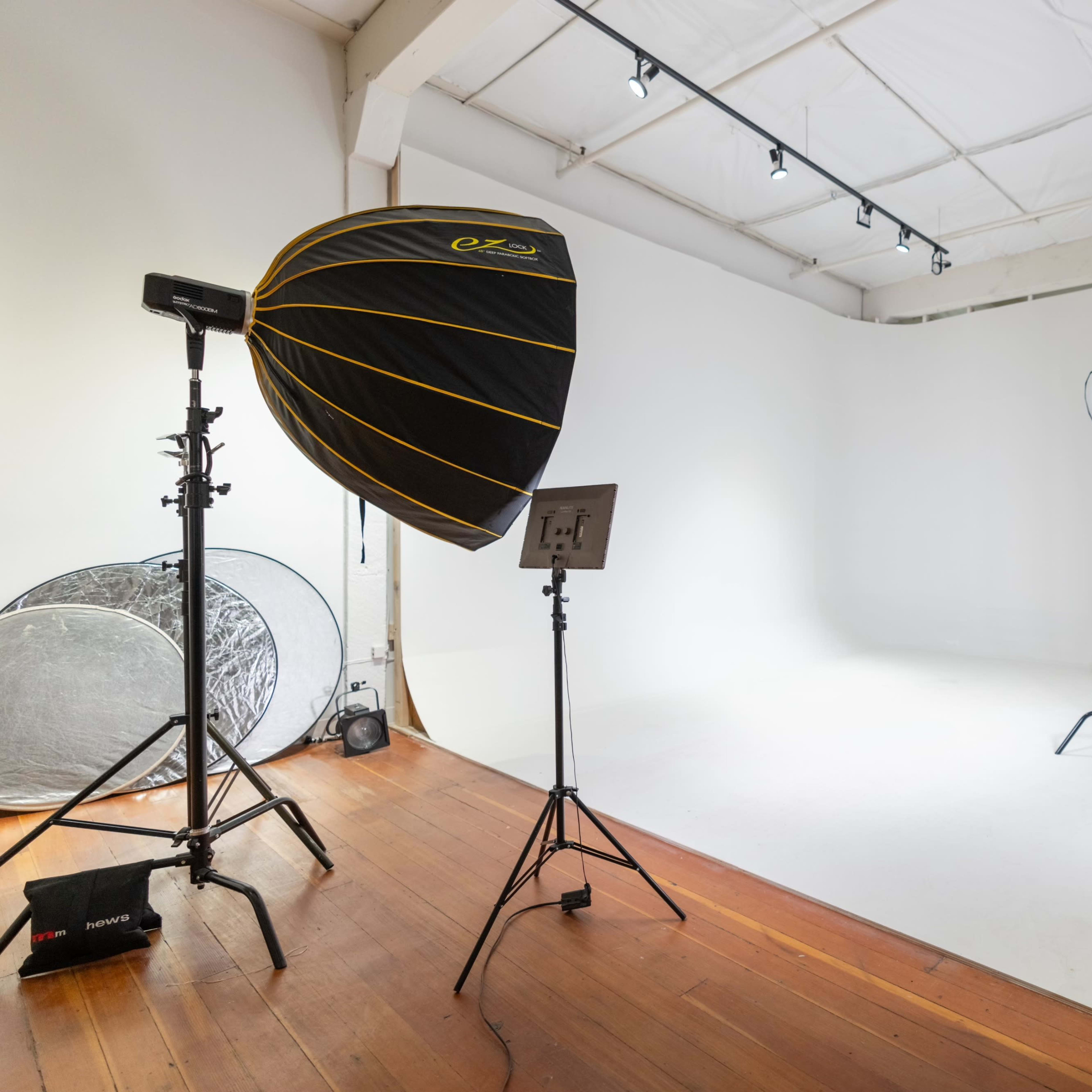 A photography studio with a white backdrop, two softbox lights, a reflector, and wooden flooring.