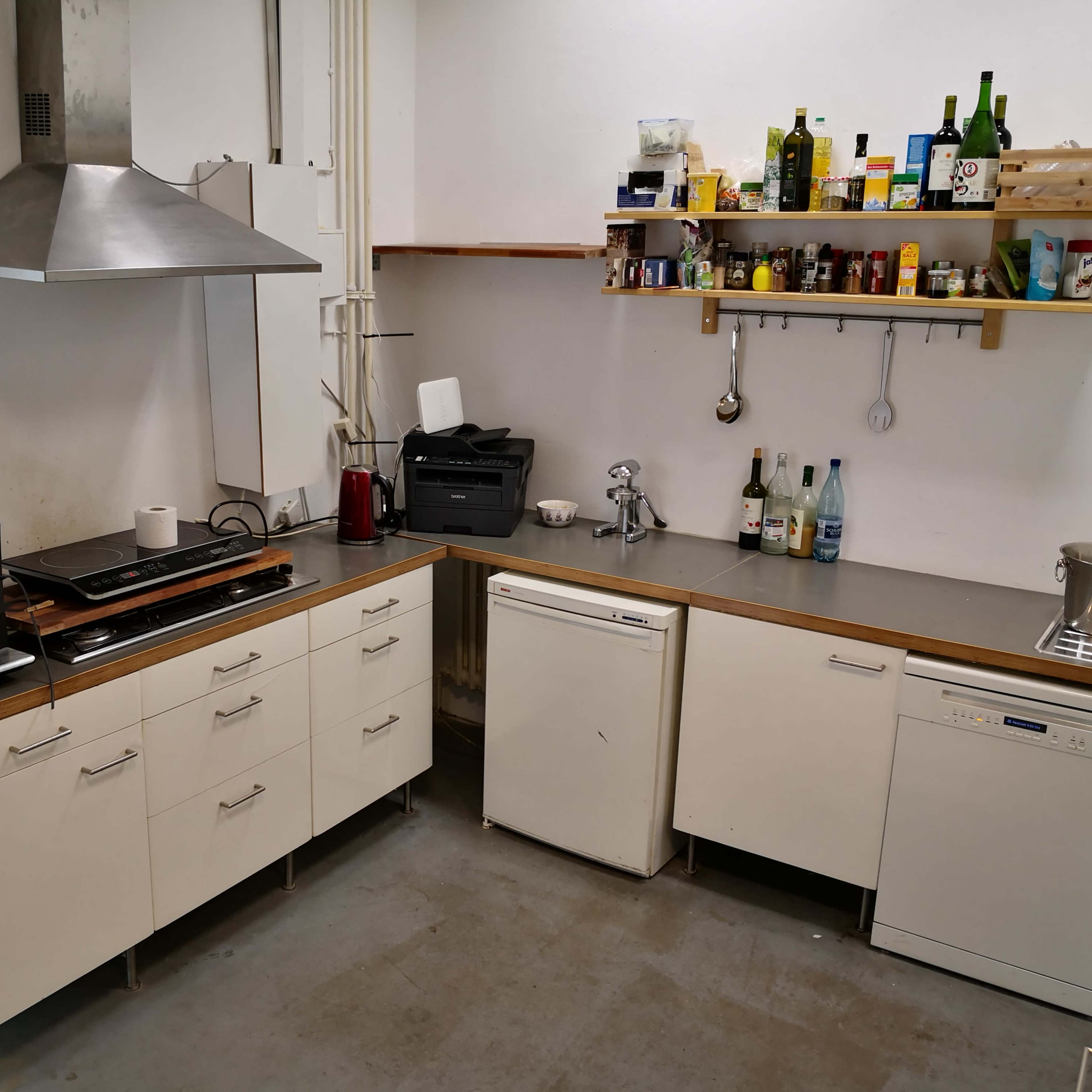 A kitchen with a stainless steel hood, a countertop with a stovetop, a microwave, a dishwasher, and shelves holding various kitchen items and bottles.