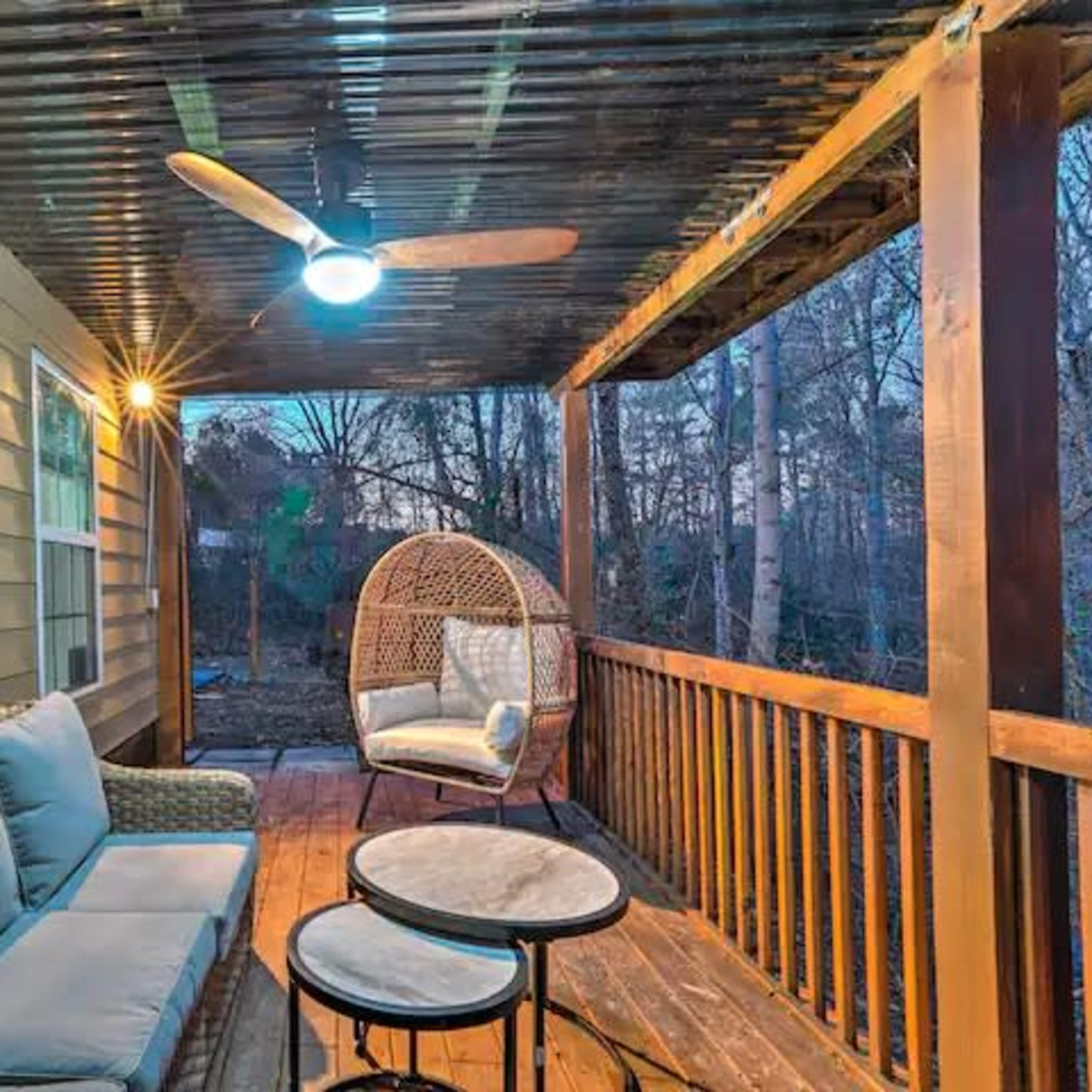 A porch with a seating area featuring a sofa, a round table, and a hanging chair, beneath a roof with wooden beams and a ceiling fan.