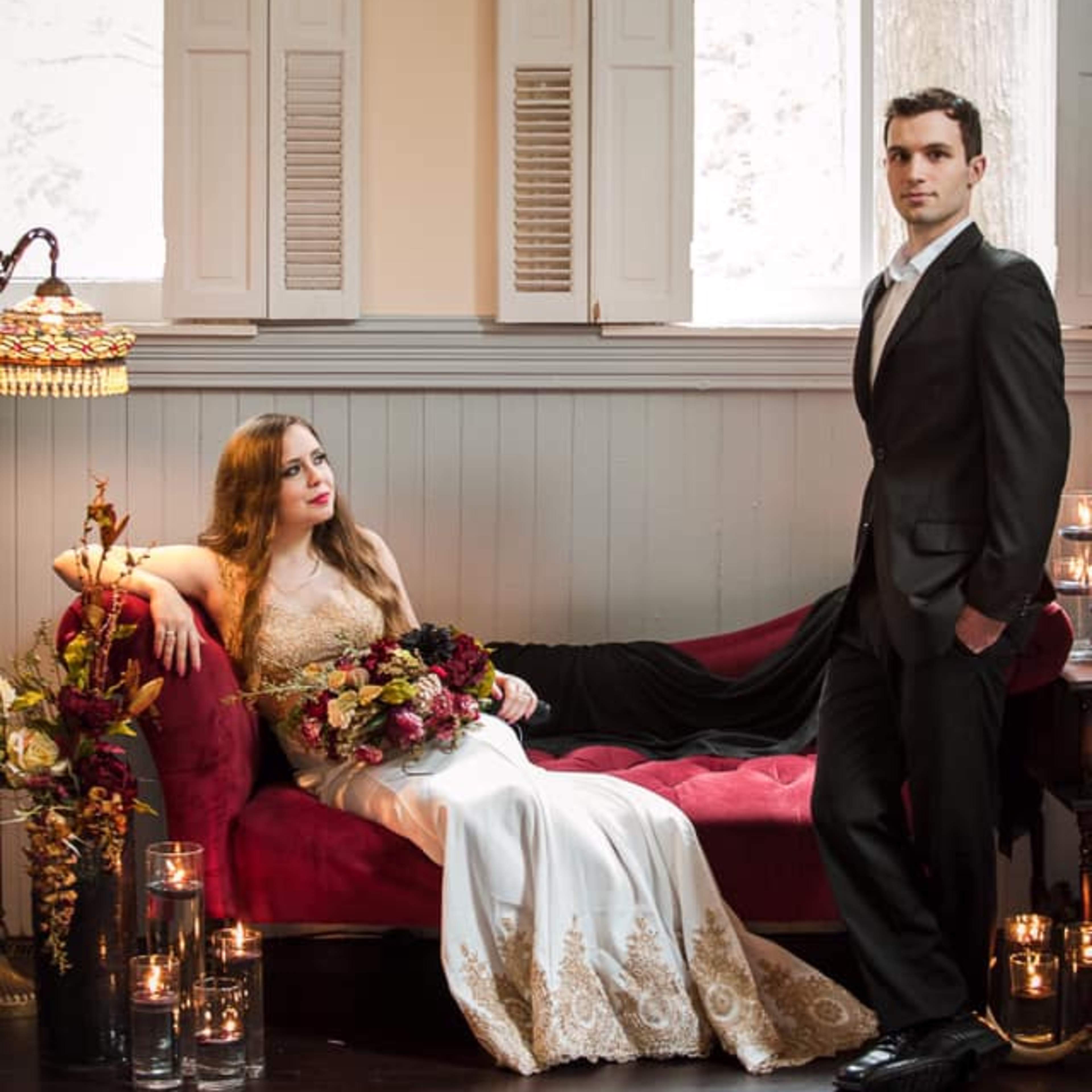 A bride in an elegant dress reclines on a red couch while a groom stands beside her in a black suit, surrounded by candles and floral arrangements.