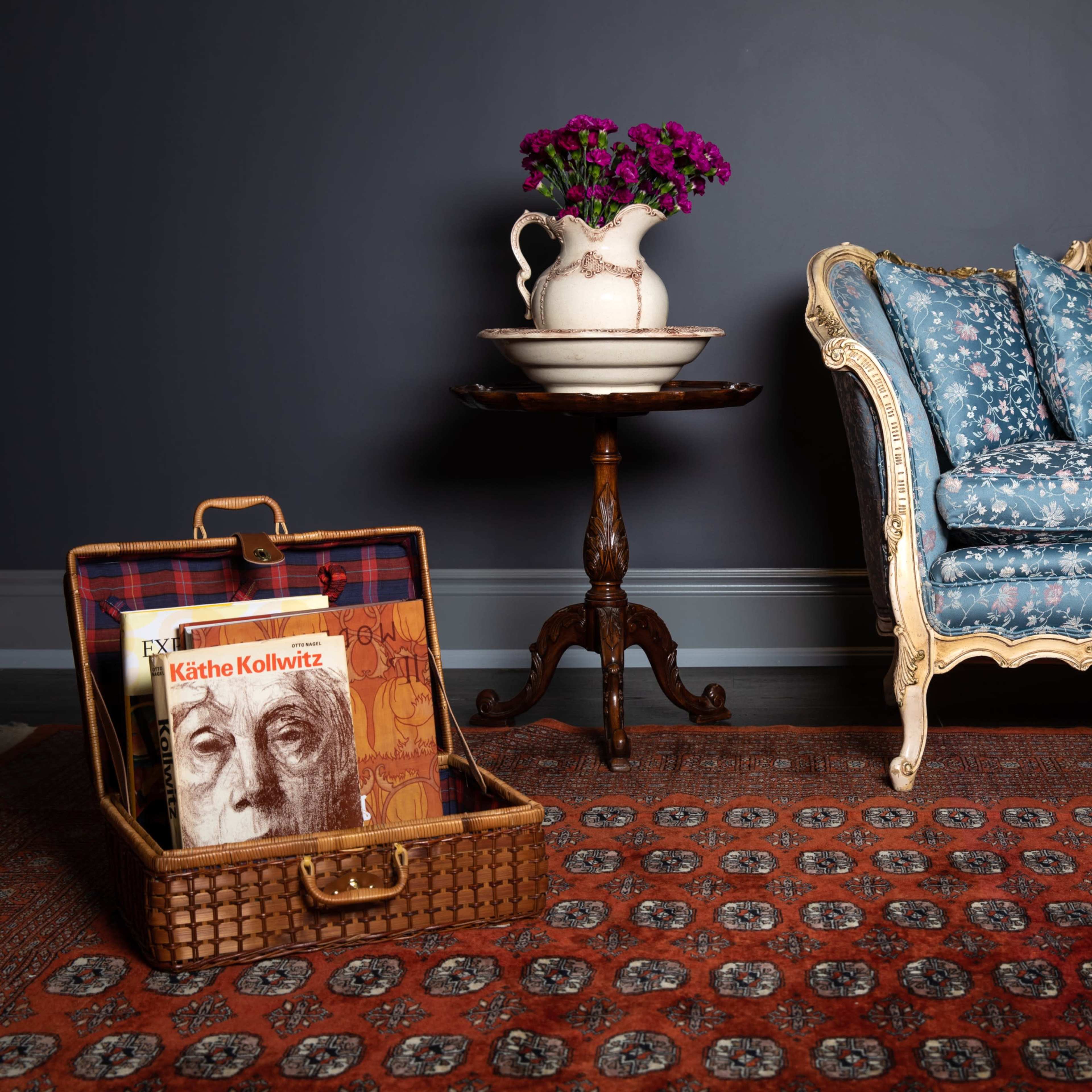 A vintage loveseat with floral upholstery is positioned next to a small table featuring a vase of flowers and a wicker suitcase containing books.