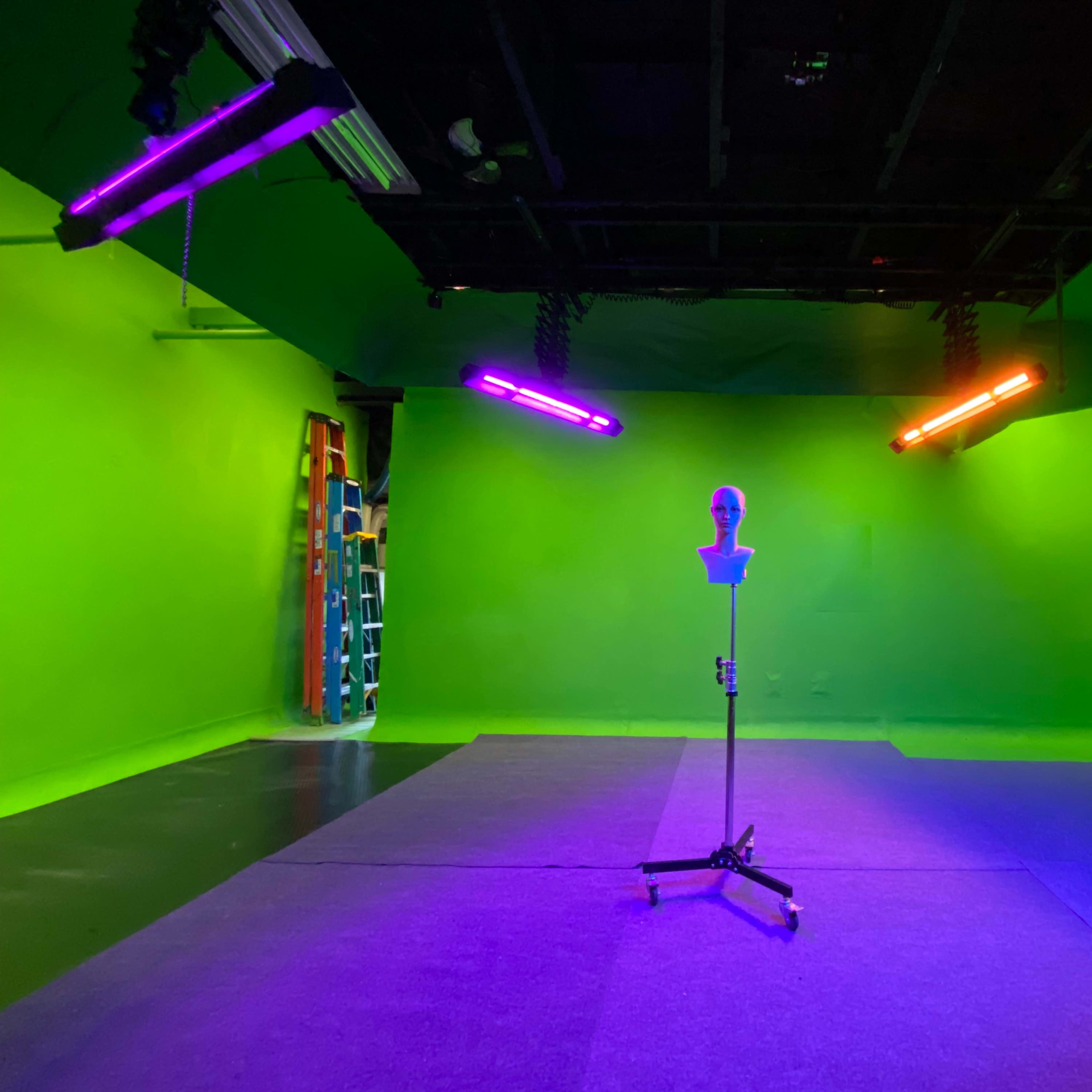 A green screen studio with a mannequin head on a stand in the center, illuminated by colored lights from above.