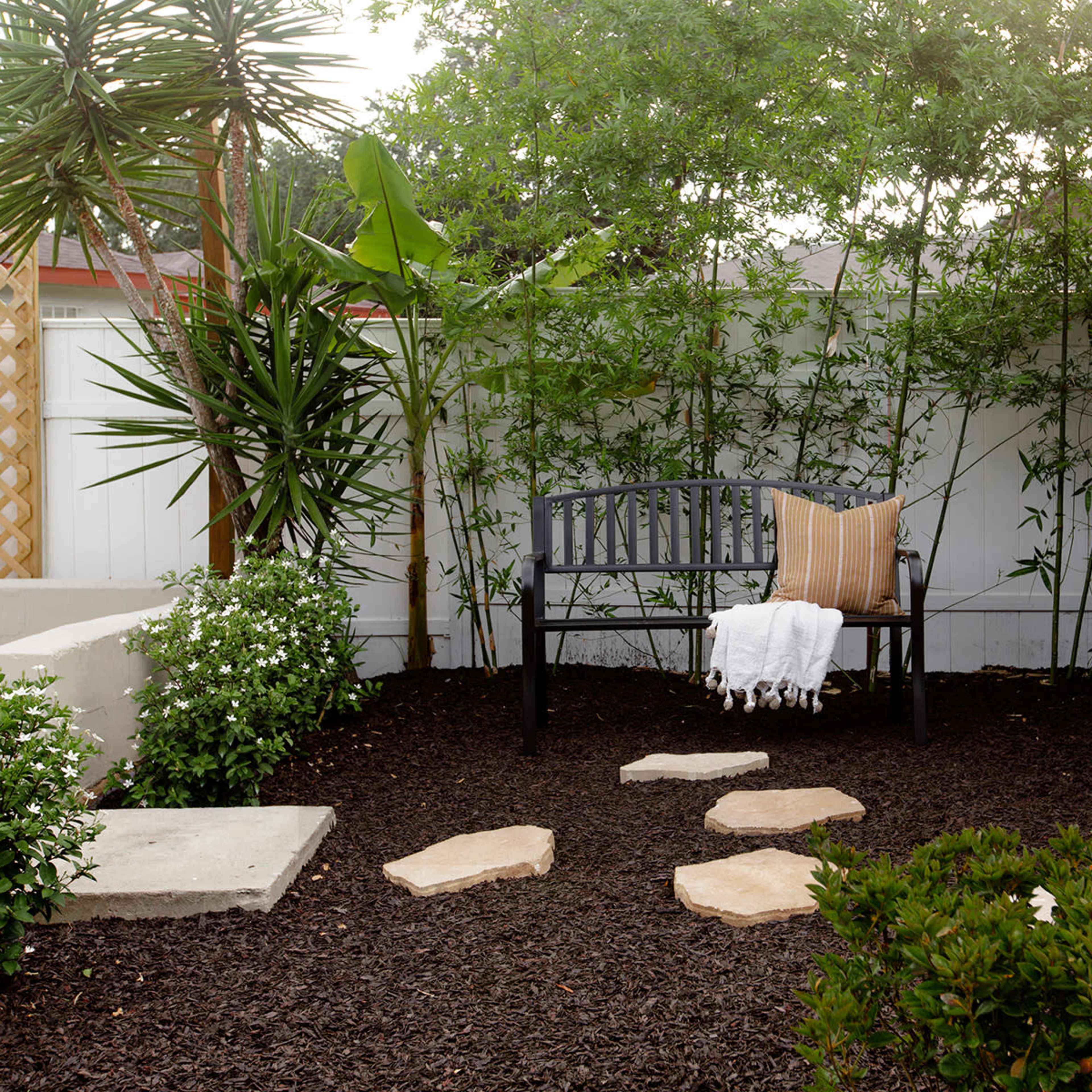 A wooden bench is placed on dark mulch in a small garden, with stone stepping paths and various plants surrounding it.