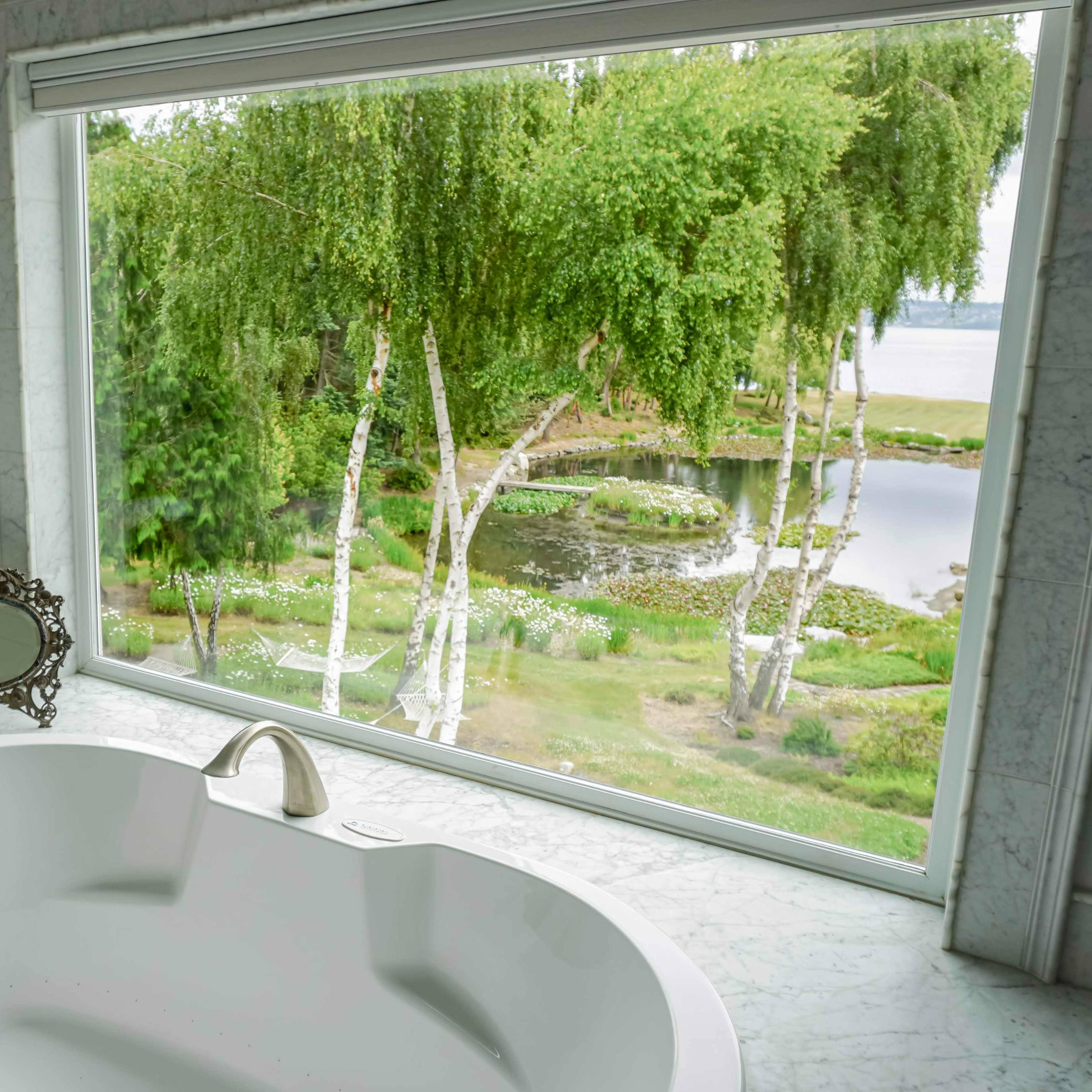 A modern bathroom features a large tub in front of a window overlooking a lush landscape with a pond and trees.