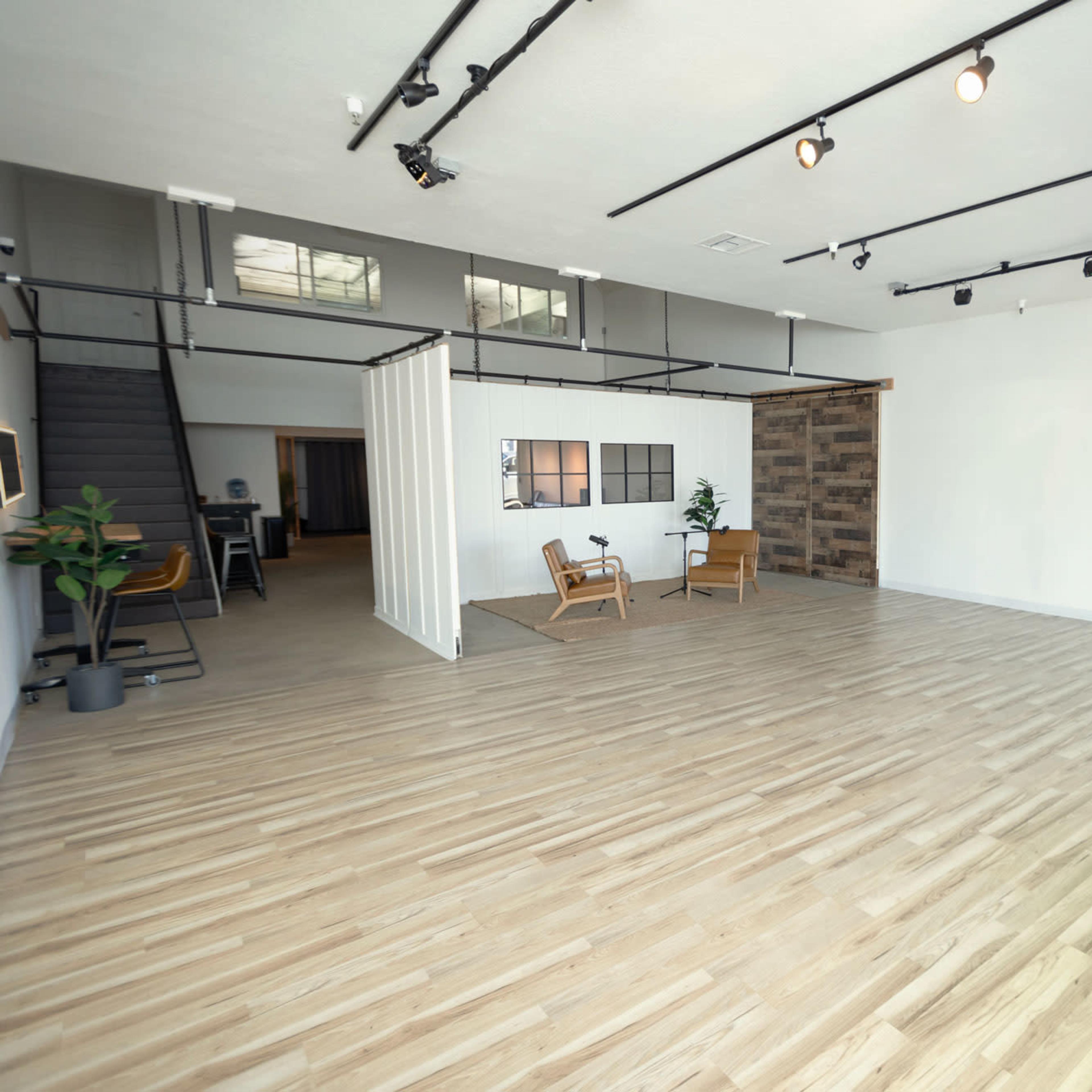 A spacious, modern room with wooden flooring, minimal furniture, and large windows allowing natural light to enter.