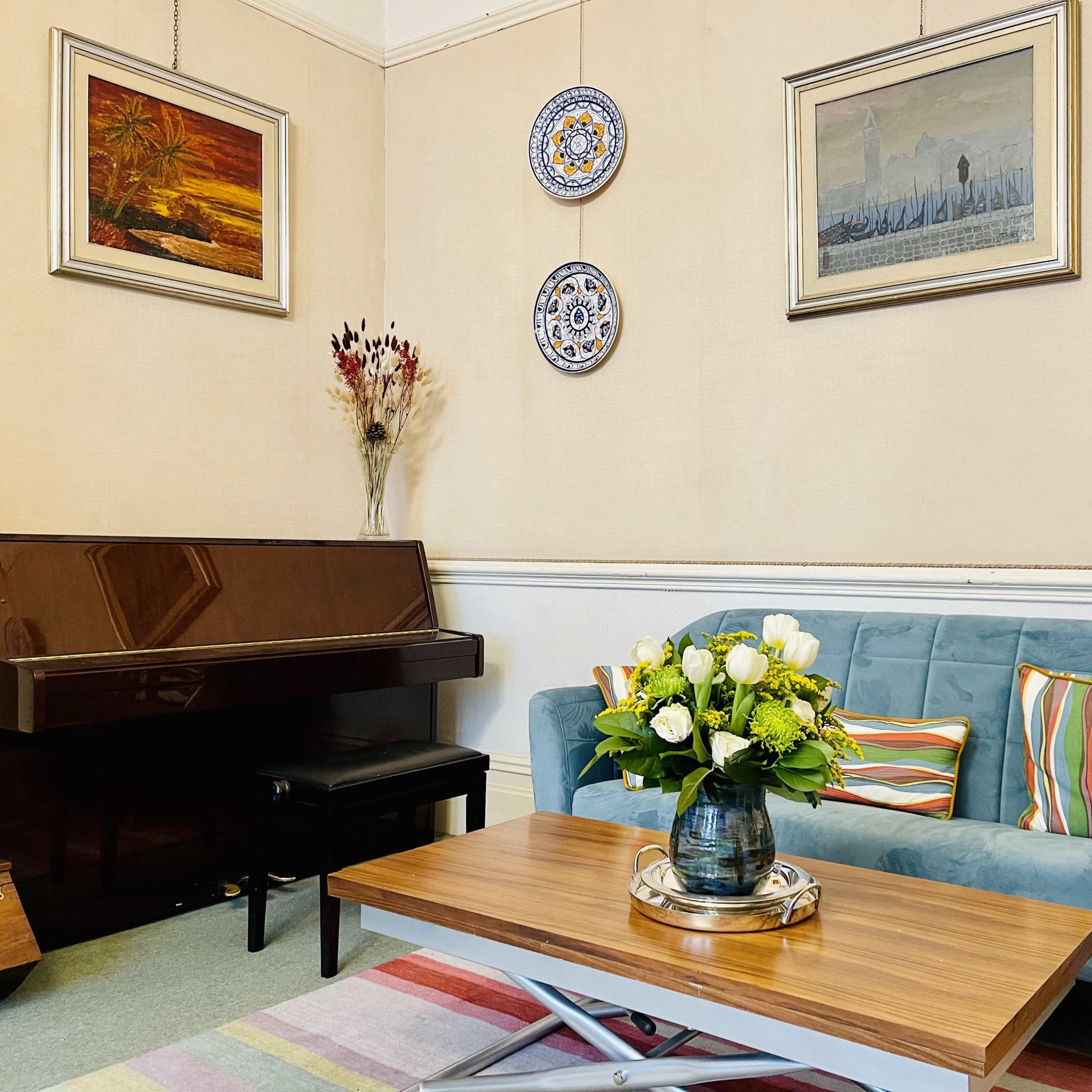 The scene features a living room with a blue sofa, a wooden coffee table, a piano, and two framed artworks on the walls, complemented by decorative plates and a vase of flowers on the table.