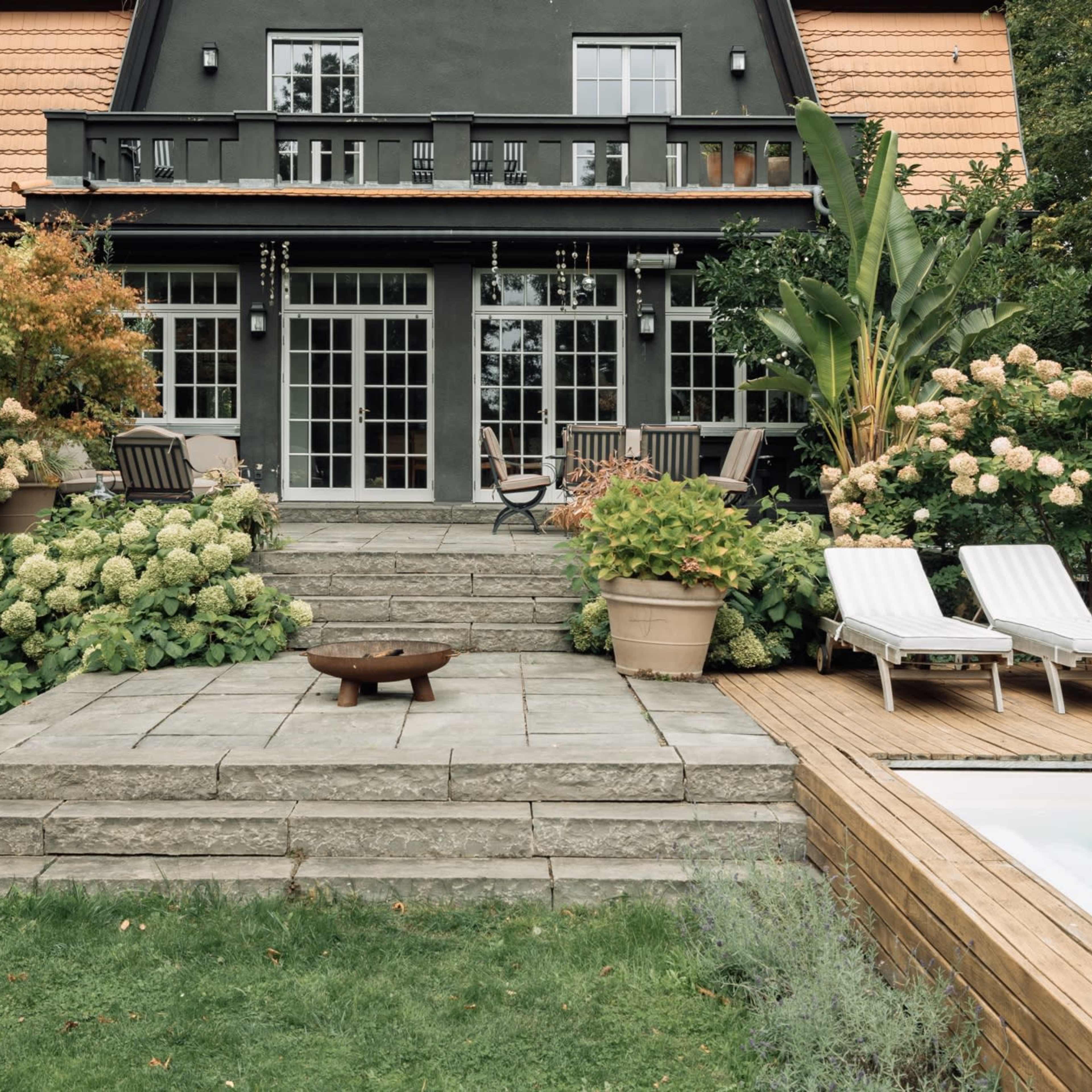 A well-landscaped backyard features a gray stone patio with a fire pit, surrounded by lush greenery and flowering hydrangeas, adjacent to a swimming pool and a two-story house with a black exterior and orange roof.